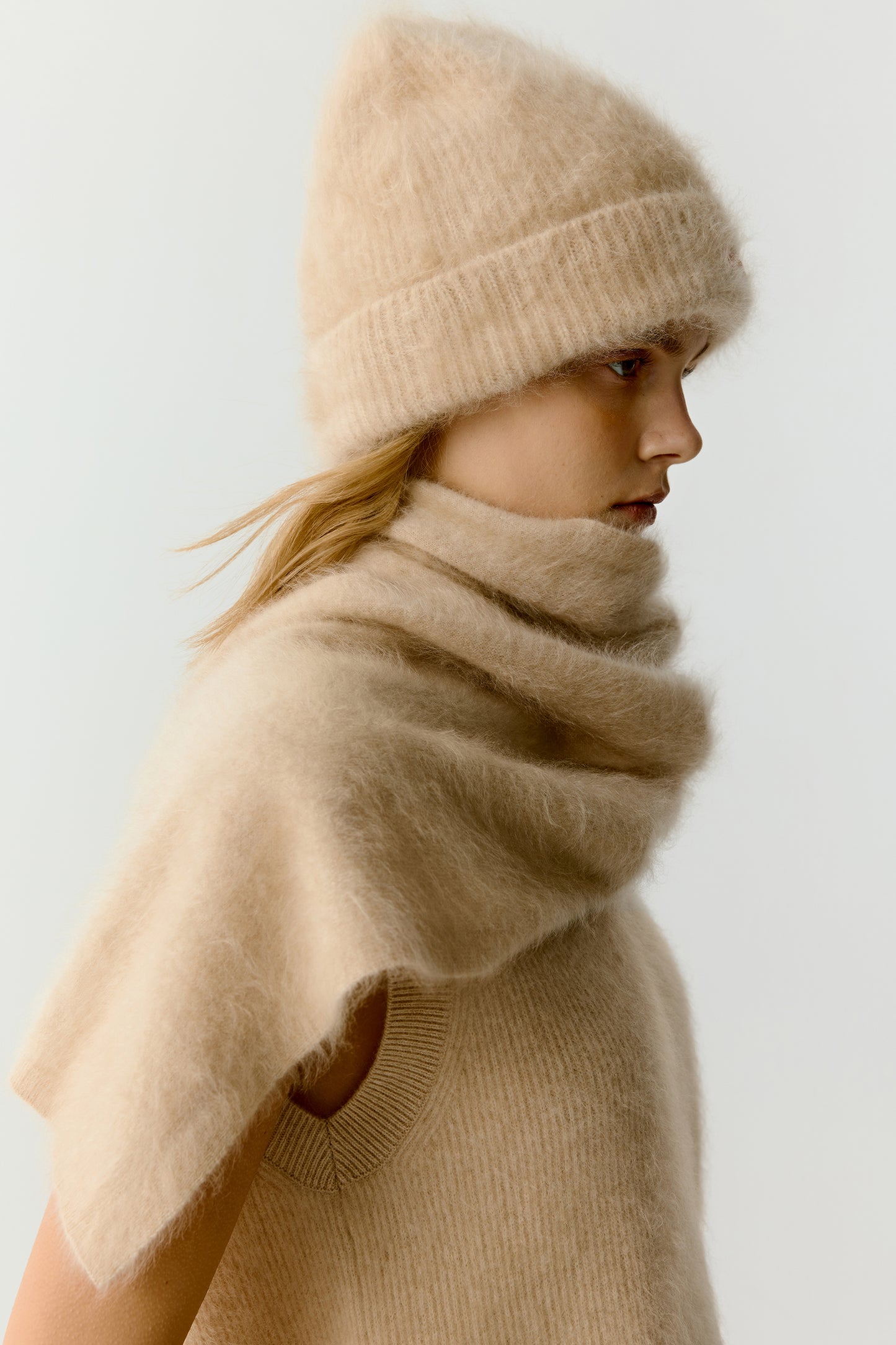 Side view of a woman wearing a beige knit beanie and scarf on a light background