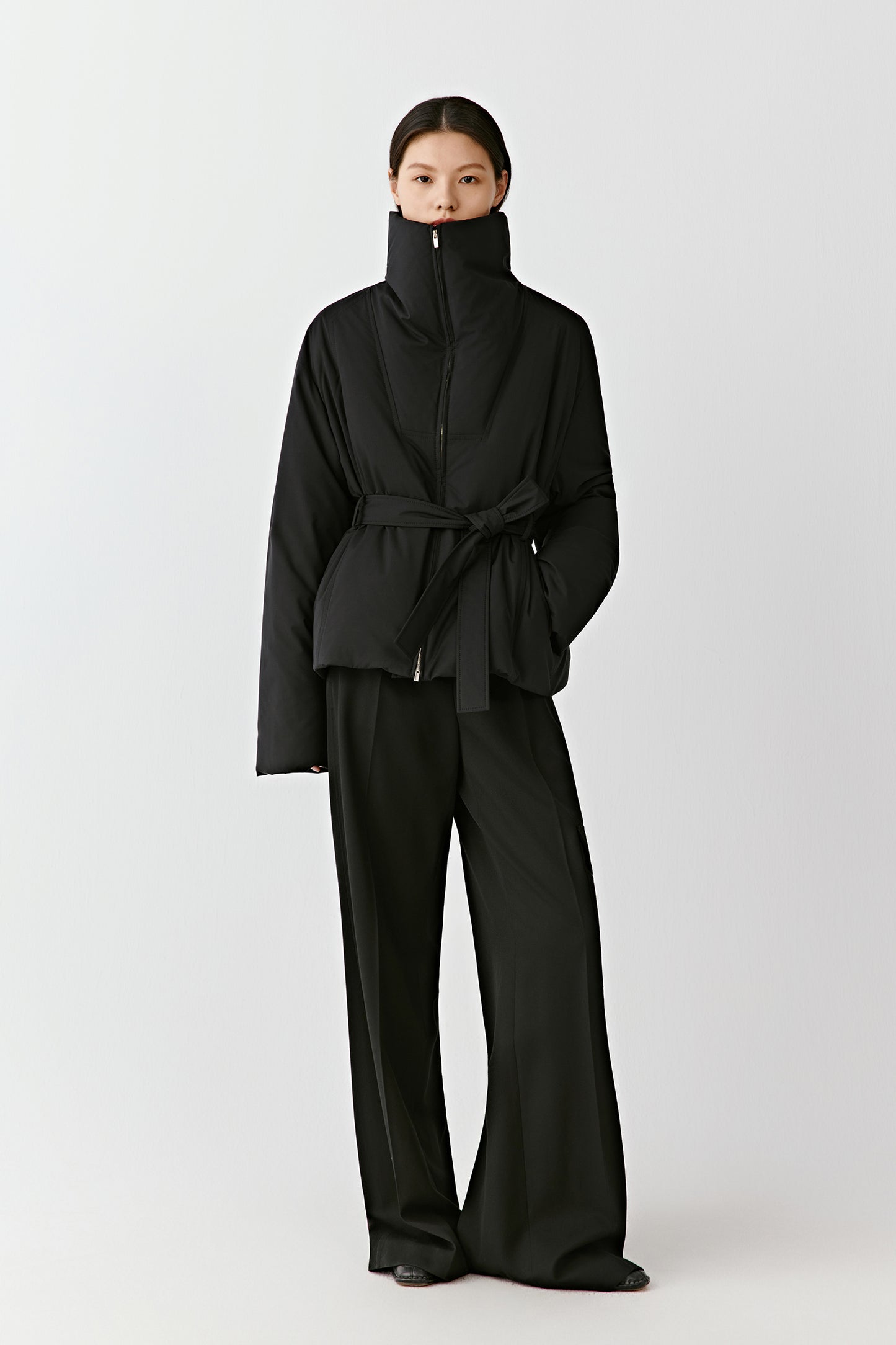 Front view of a woman wearing a black wool and silk DEW blouson with a high stand collar and belted waist.