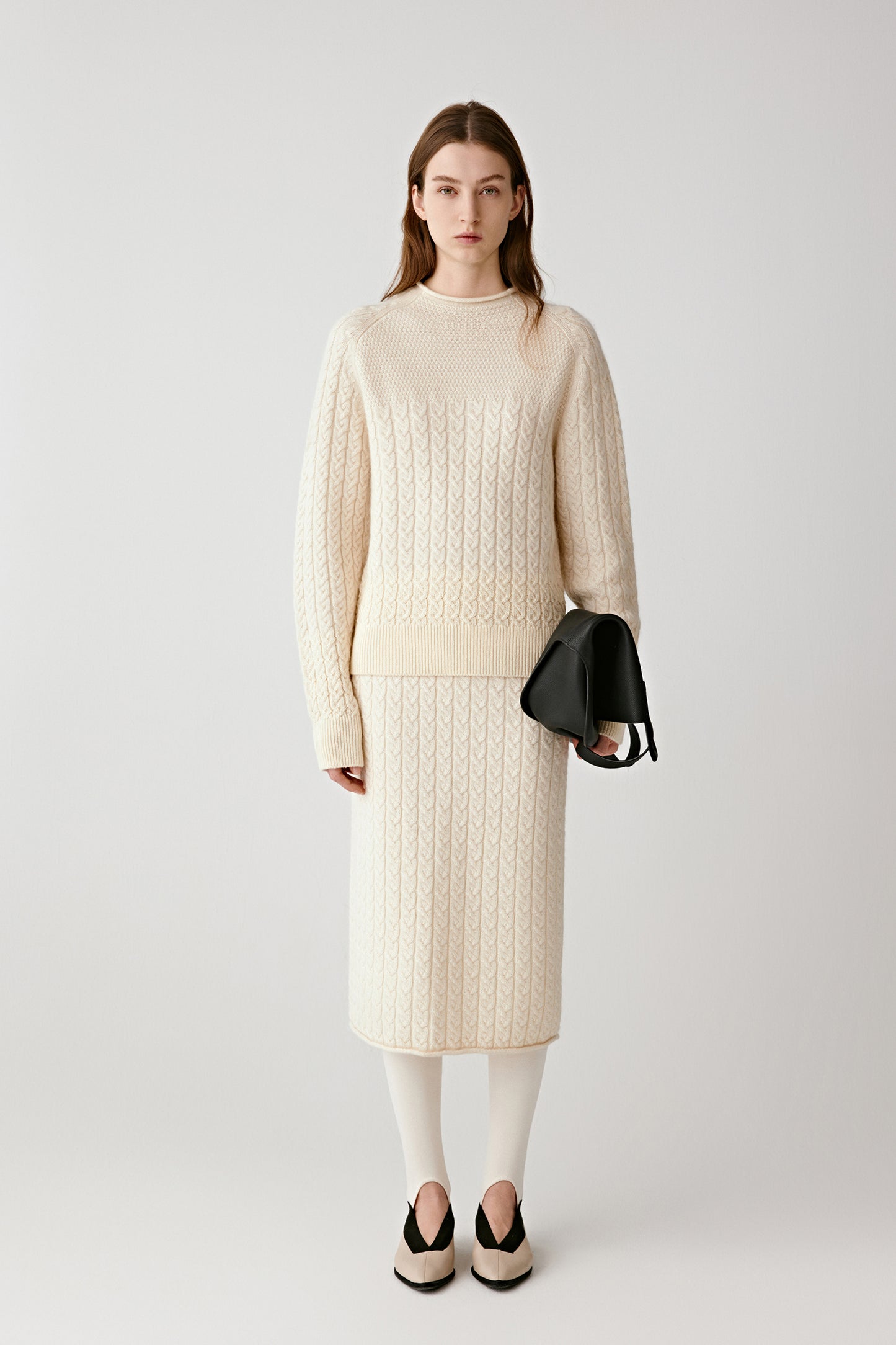 Woman wearing a ivory cable knit sweater and skirt set with a black handbag on a white background