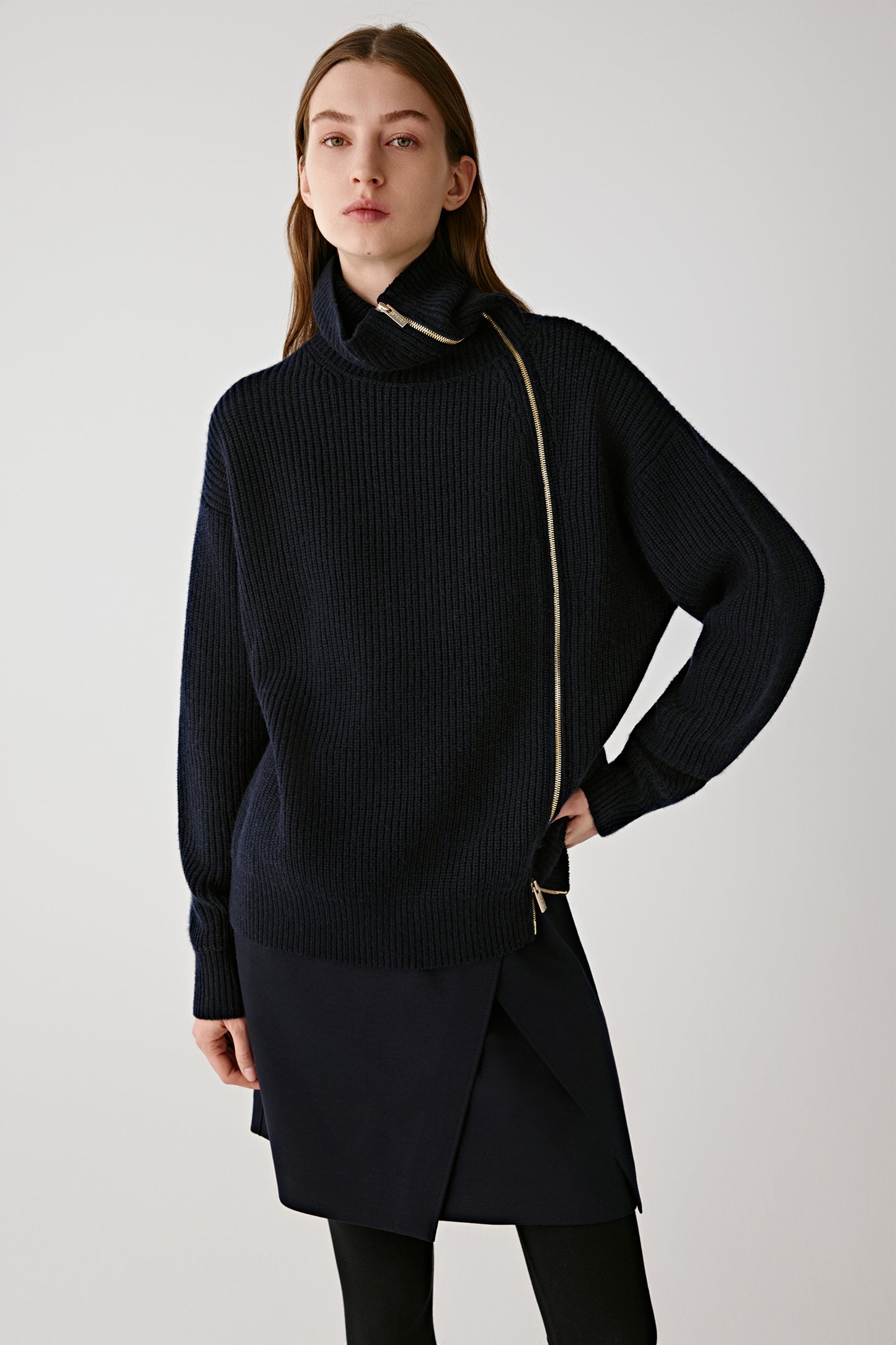 Front view of a model wearing a navy blue asymmetrical wool and cashmere cardigan with a diagonal gold zip fastening and ribbed texture.