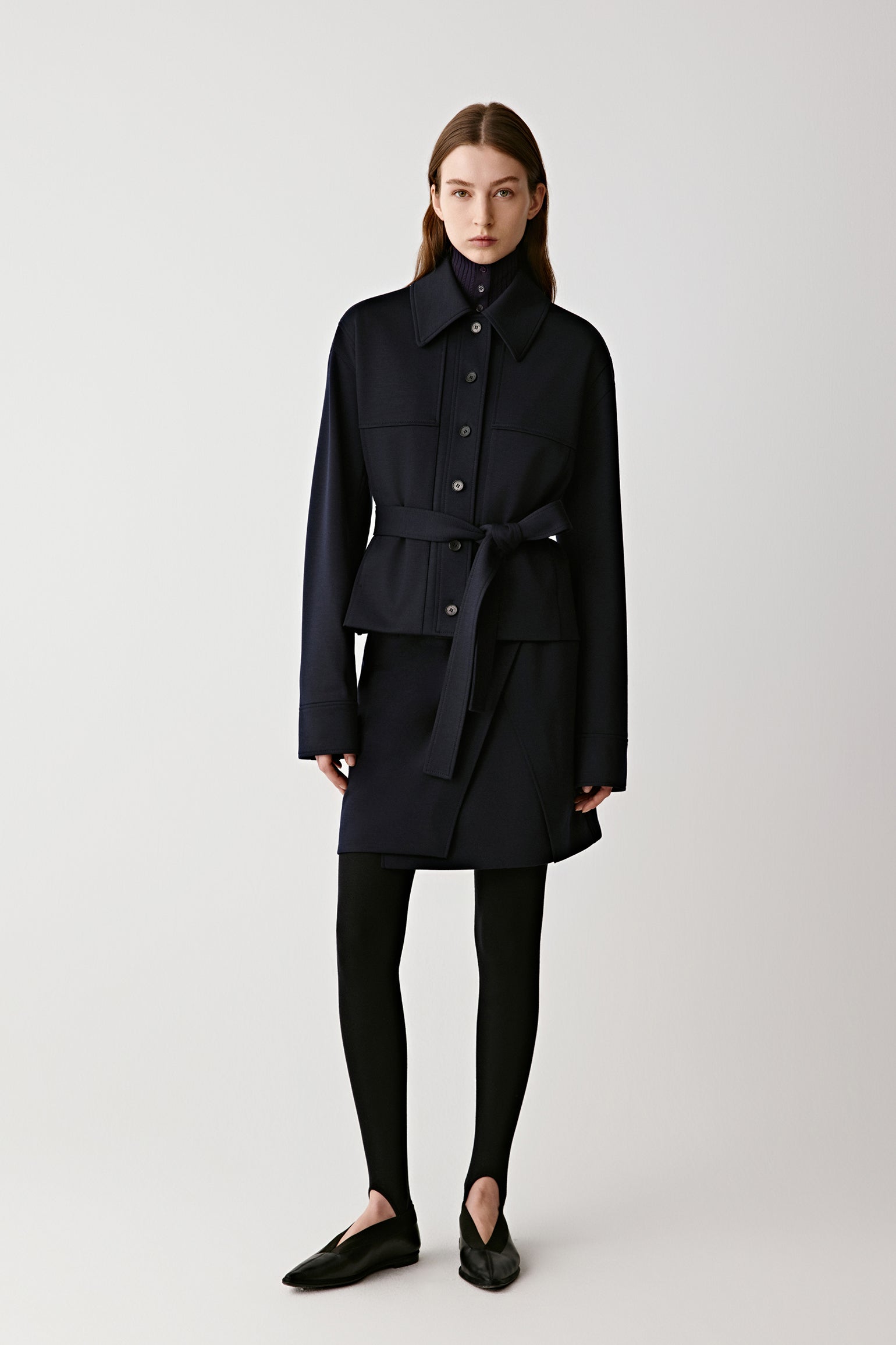 Full-length front view of the model wearing the navy wool jacket with a belted waist, paired with black trousers, showing its structured, timeless silhouette.