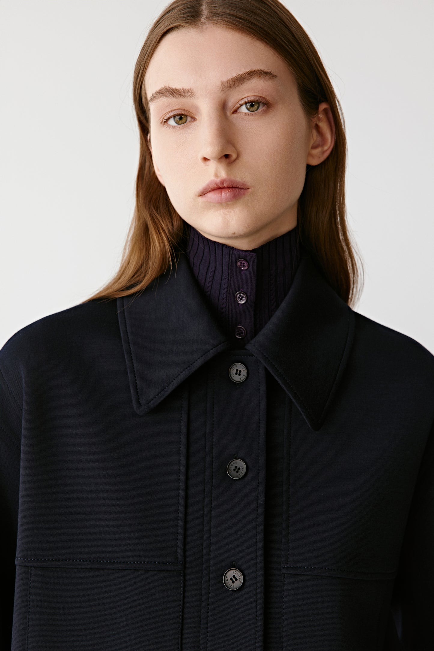 Close-up portrait of the model wearing the navy worsted wool jacket. The image highlights the structured collar, front button placket, and the texture of the fabric. A dark ribbed turtleneck in a deep purple tone is layered underneath, adding contrast and sophistication. The lighting emphasizes the clean tailoring and refined materials.