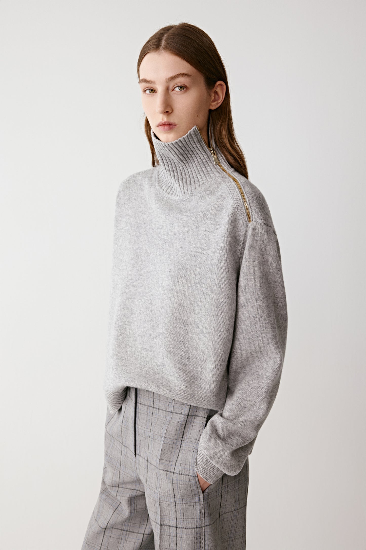 Full-length front view of the model wearing the light grey cashmere jumper with a ribbed funnel neck and zip detail, paired with matching trousers.