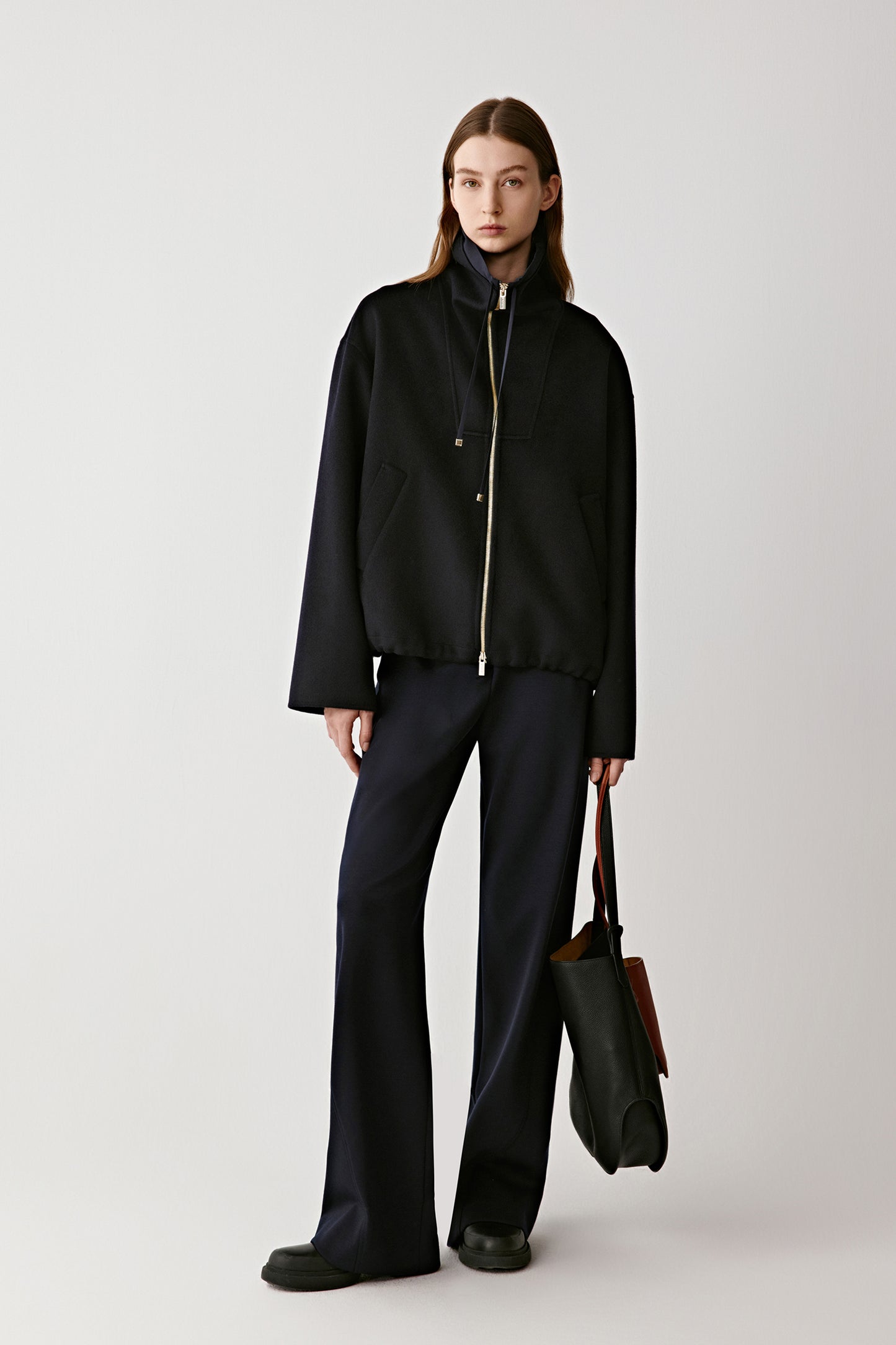 Front view of a model wearing a black double-faced wool and silk zip jacket, styled with wide-leg trousers and a sleek handbag.