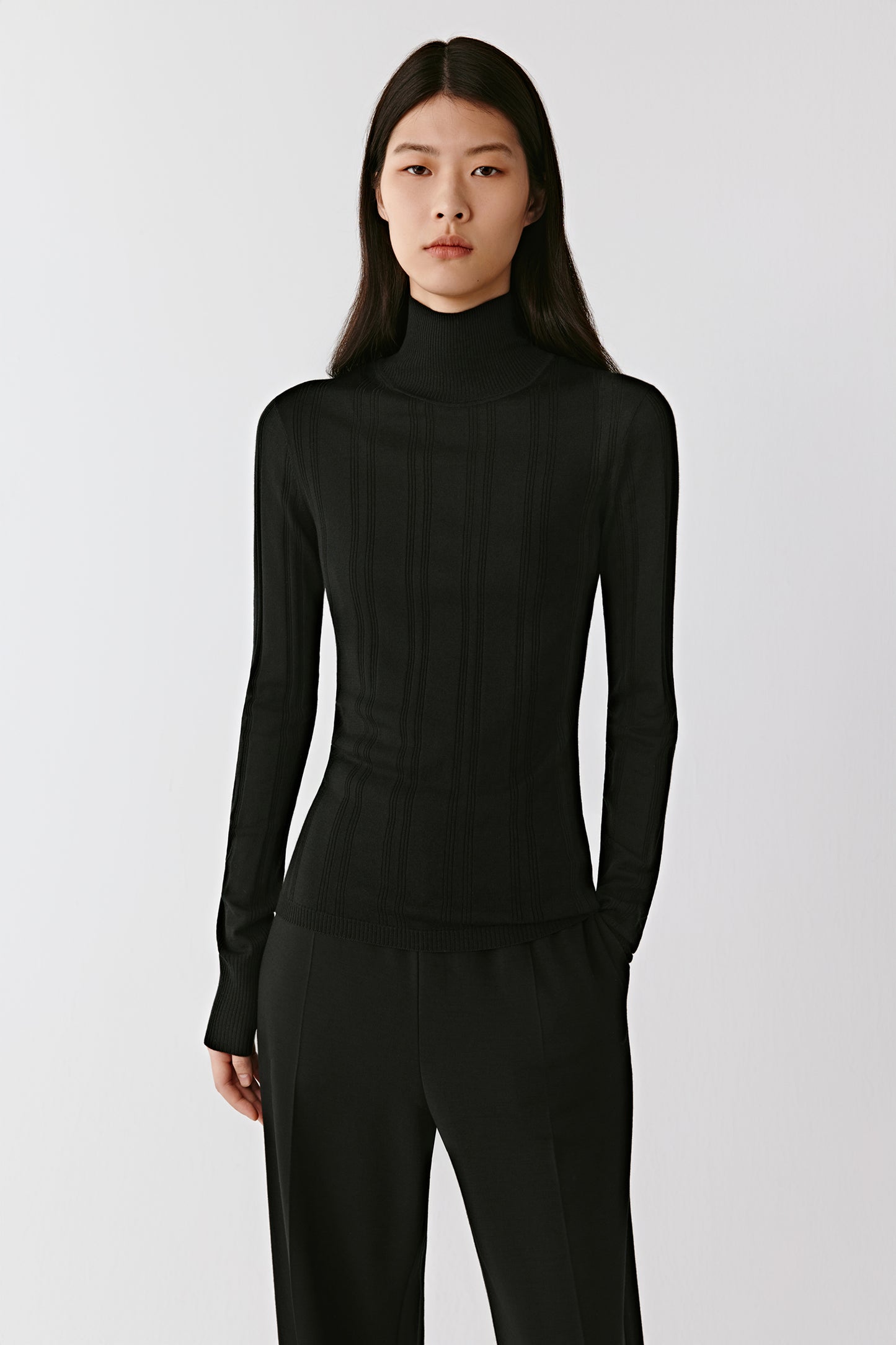A woman stands facing forward wearing a black ribbed mock-neck pullover in wool and silk.