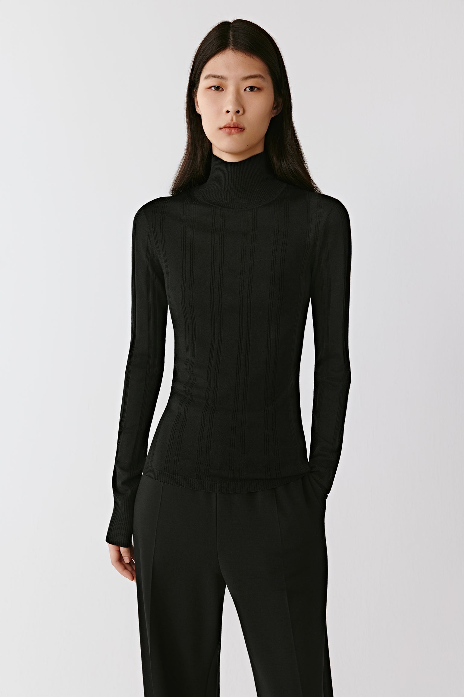A woman stands facing forward wearing a black ribbed mock-neck pullover in wool and silk.