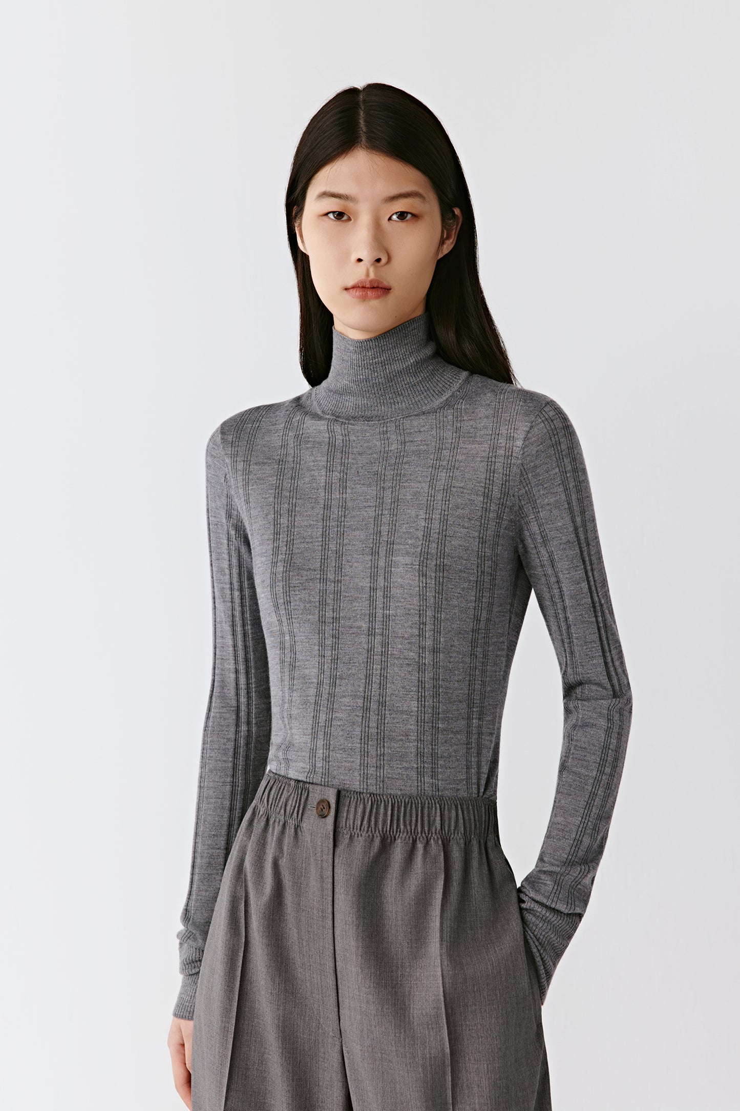 A woman stands facing forward wearing a smoky grey ribbed mock-neck pullover in wool and silk.