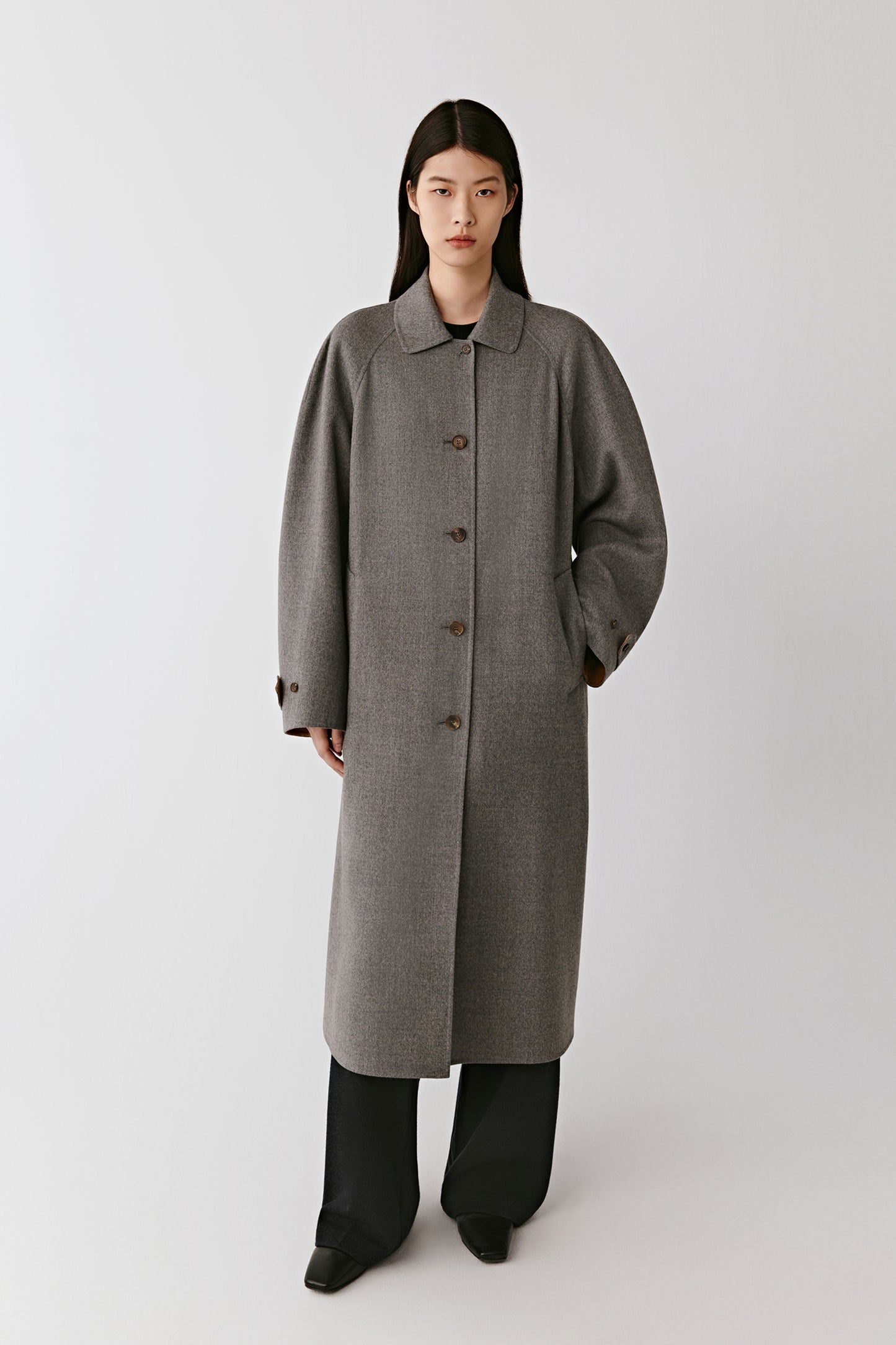 A woman wearing a long grey double-faced wool coat with a relaxed, straight silhouette and black trousers. The clean lines and classic collar highlight the minimalist tailoring.