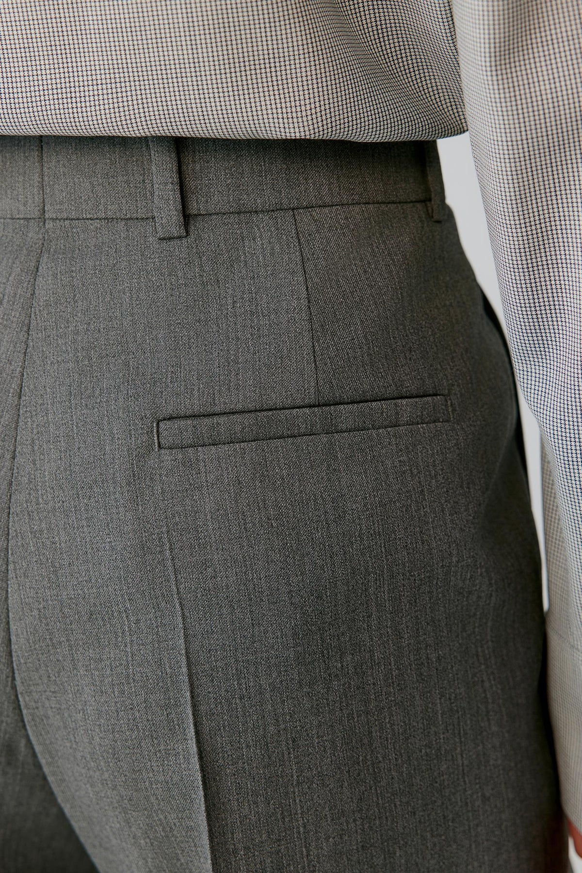Close-up of dark gray tailored trousers with a welt pocket and belt loop, shown on a model wearing a small check shirt.