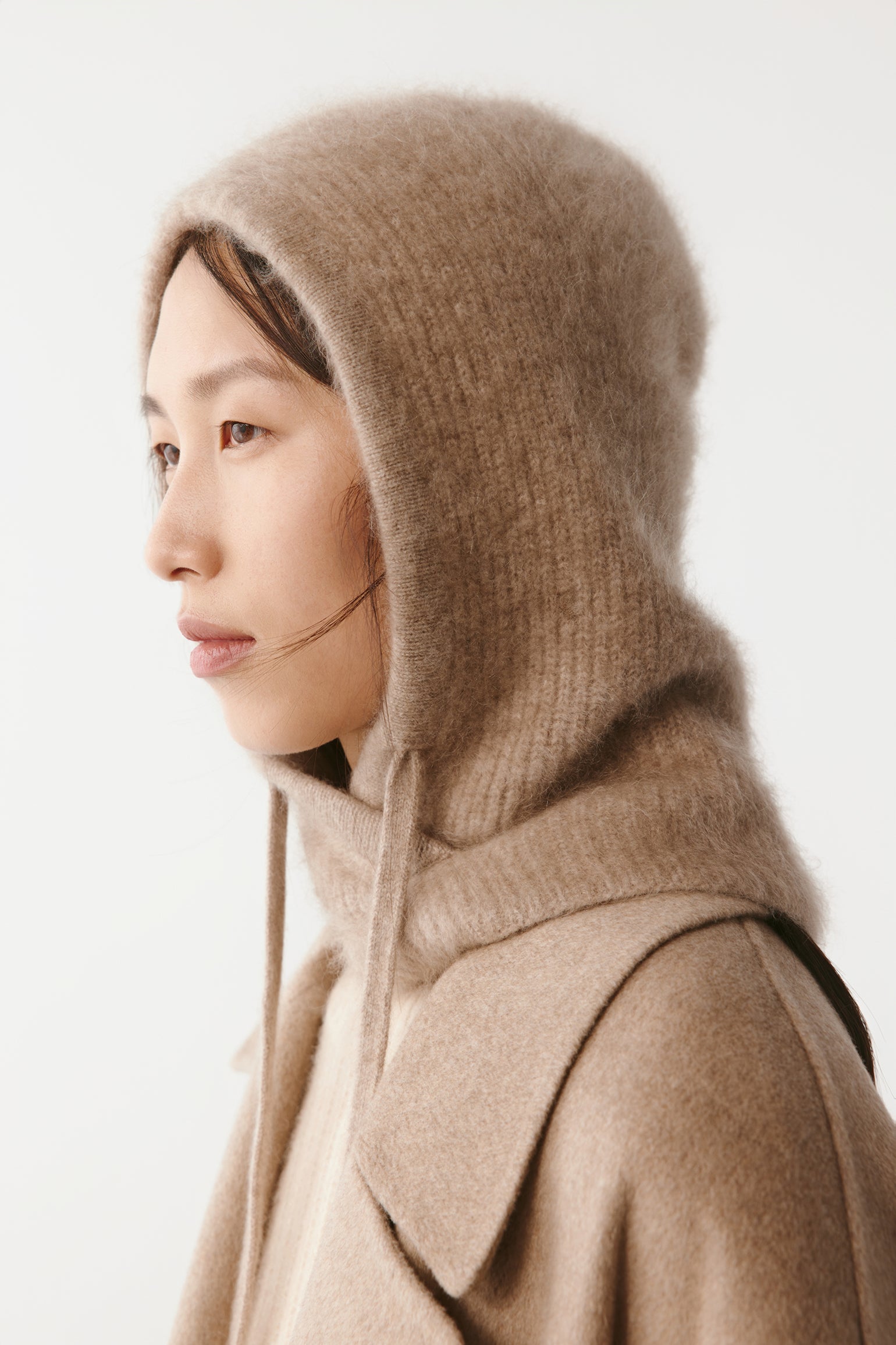 Side view of a woman wearing a beige knit hood with long ties on a light background