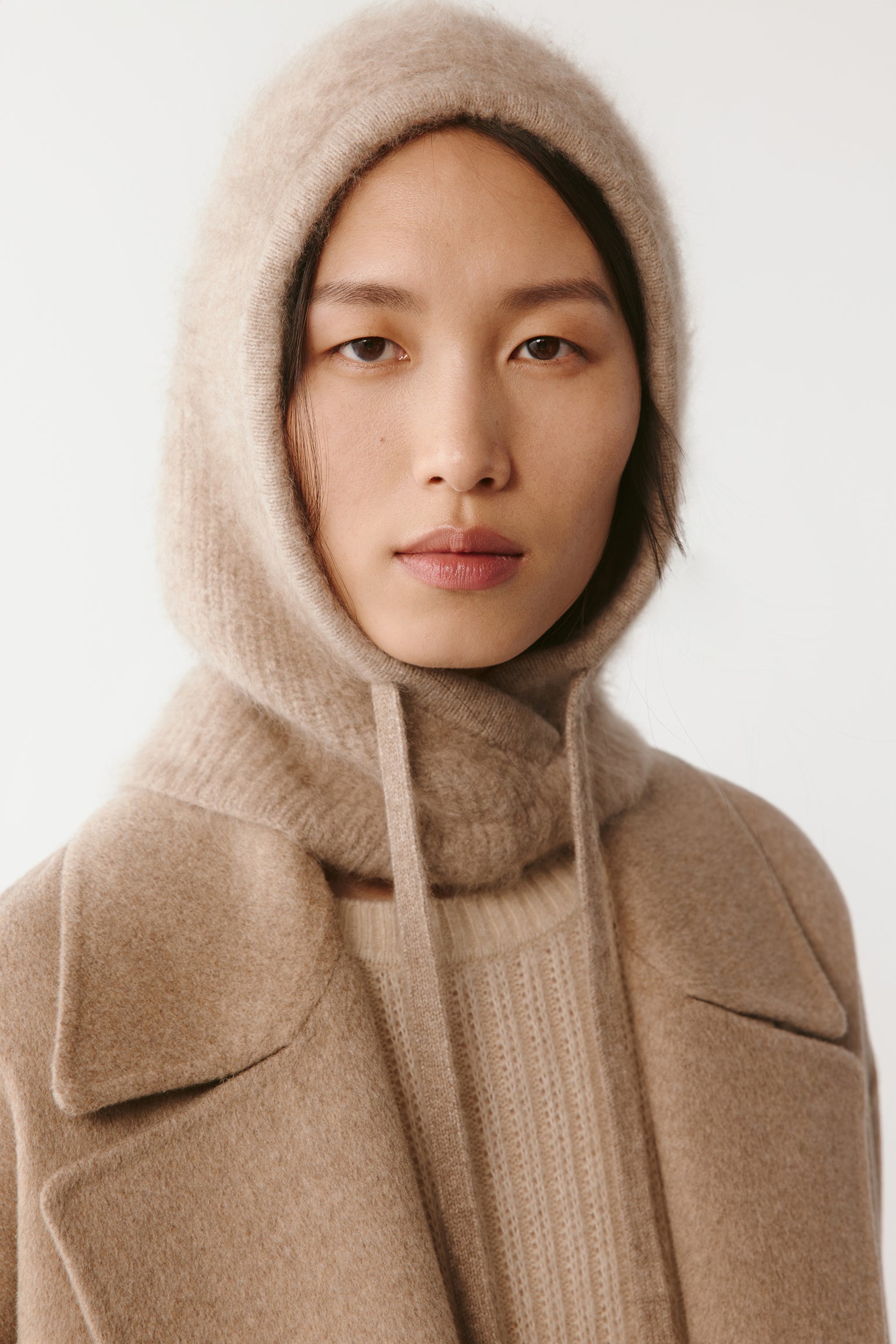 Woman wearing a beige knit hood with long drawstrings styled with a beige coat on a light background
