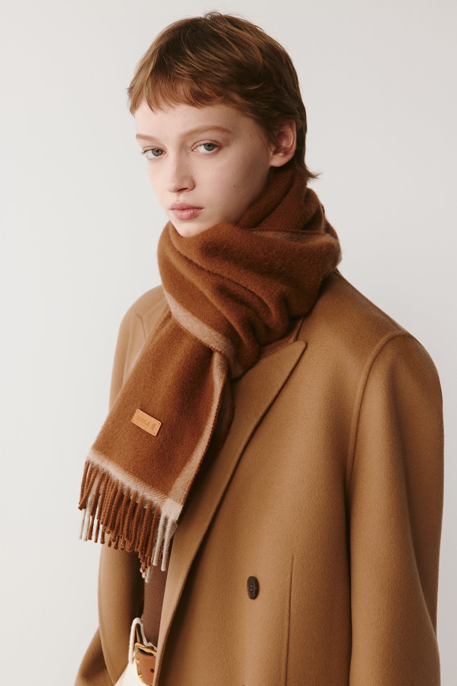 Woman wearing a brown wool scarf with fringed edges styled with a camel coat on a light background