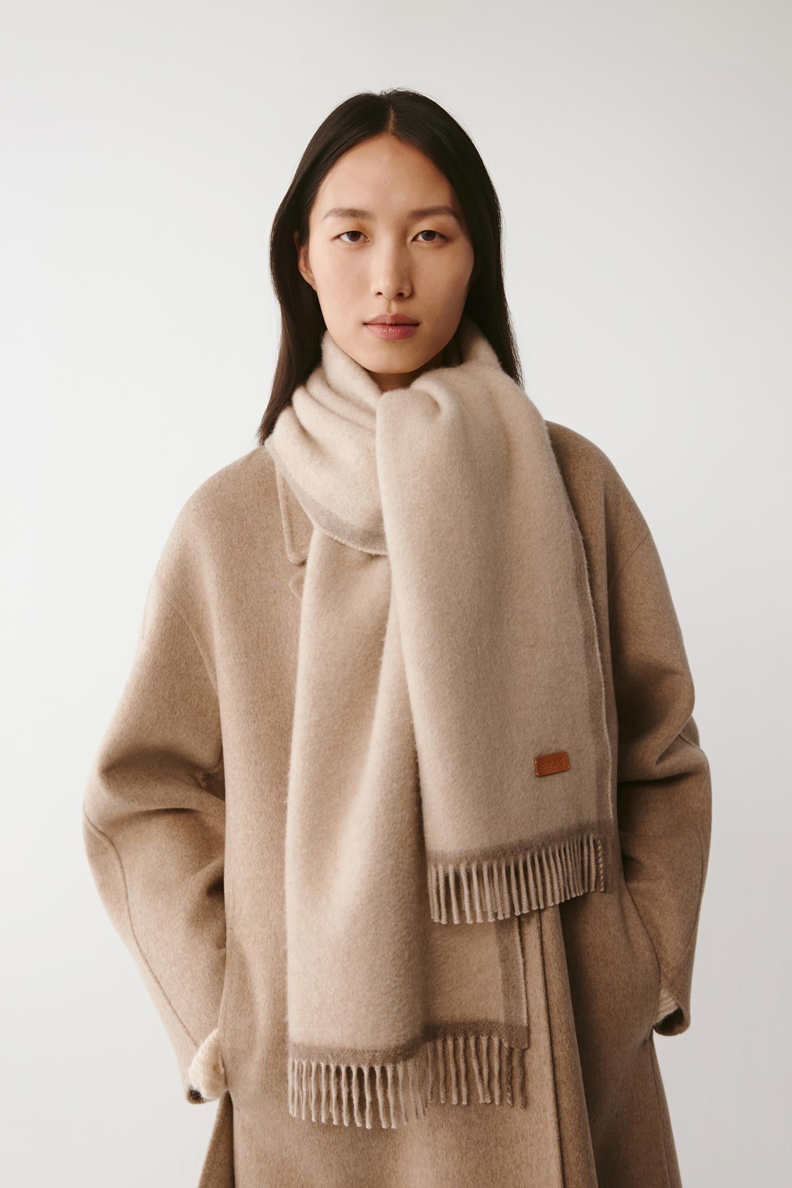 Woman wearing a large cream wool scarf styled with a beige coat on a light background