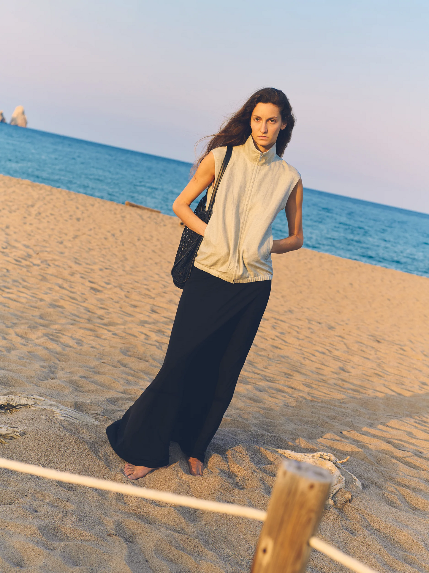 Female model standing barefoot on a sandy beach at golden hour, looking back over her shoulder — wearing a sleeveless zip-up gilet in soft ivory linen with a high funnel neck, structured shoulders and a cropped, fitted silhouette, paired with a fluid dark navy maxi skirt in lightweight linen with a straight, floor-grazing drape, and carrying a dark woven raffia tote bag — a refined, understated coastal look against a calm turquoise sea and pale evening sky