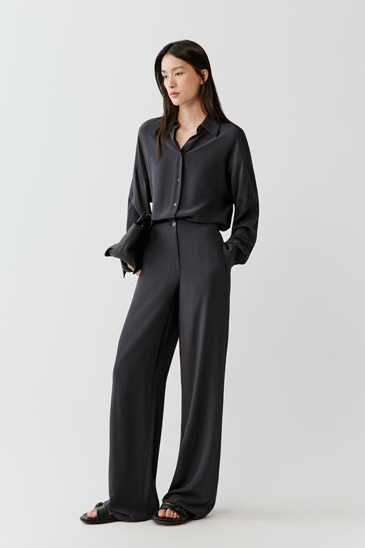 Woman wearing a black silk crepe de chine shirt with a fluid, relaxed cut, styled with matching trousers. She moves through front, side and three-quarter views, showing the lightweight fabric, soft drape and slightly matte surface. The classic collar, buttoned front and long sleeves remain visible, while the shirt falls smoothly over the body, emphasizing ease of movement and a relaxed silhouette.
