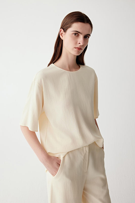Video showing a woman wearing an off-white silk blouse with a softly crinkled surface and fluid drape, moving to reveal the lightweight texture and continuous silhouette. The fabric falls softly through the body and sleeves, while the side slit and back slit fastening open subtly with movement. The motion highlights the matte crinkled texture, relaxed structure and fluid construction, with the lightweight silk shifting naturally around the body.
