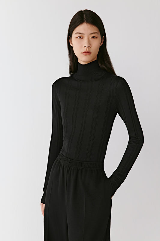 A woman wears a black ribbed pullover in wool and silk, styled with wide black trousers. She turns gracefully, allowing the camera to capture the pullover’s fluid drape and fitted shape. The soft texture of the fabric and the purity of the lines are accentuated through gentle movement. The sequence moves between full-length and close-up views, revealing the elegance and timelessness of the piece.