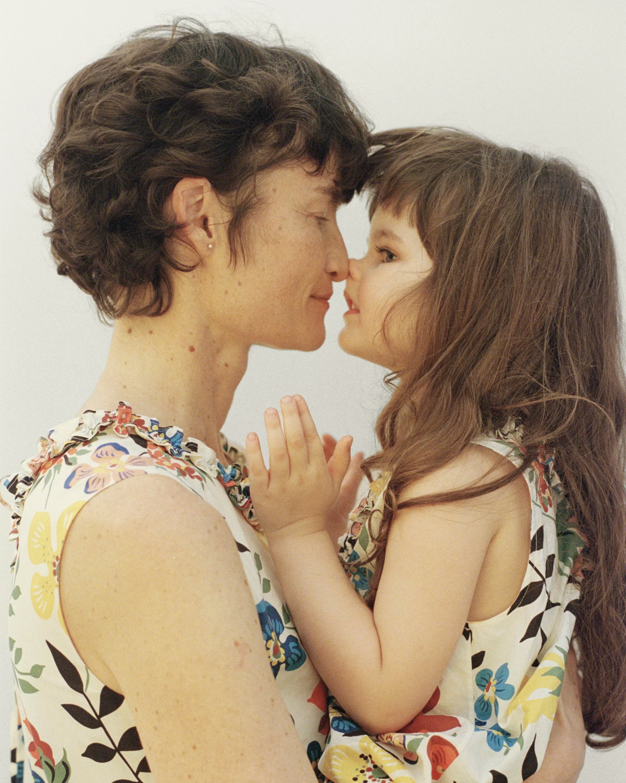 Close-up profile portrait of an adult woman and a young girl facing each other nose to nose in a tender moment, hands clasped together between them; both wearing matching sleeveless dresses in lightweight fabric with a vibrant Liberty floral print featuring blue, red and yellow blooms with dark botanical stems on an ivory ground, with delicate ruffle trim at the neckline — against a soft white background