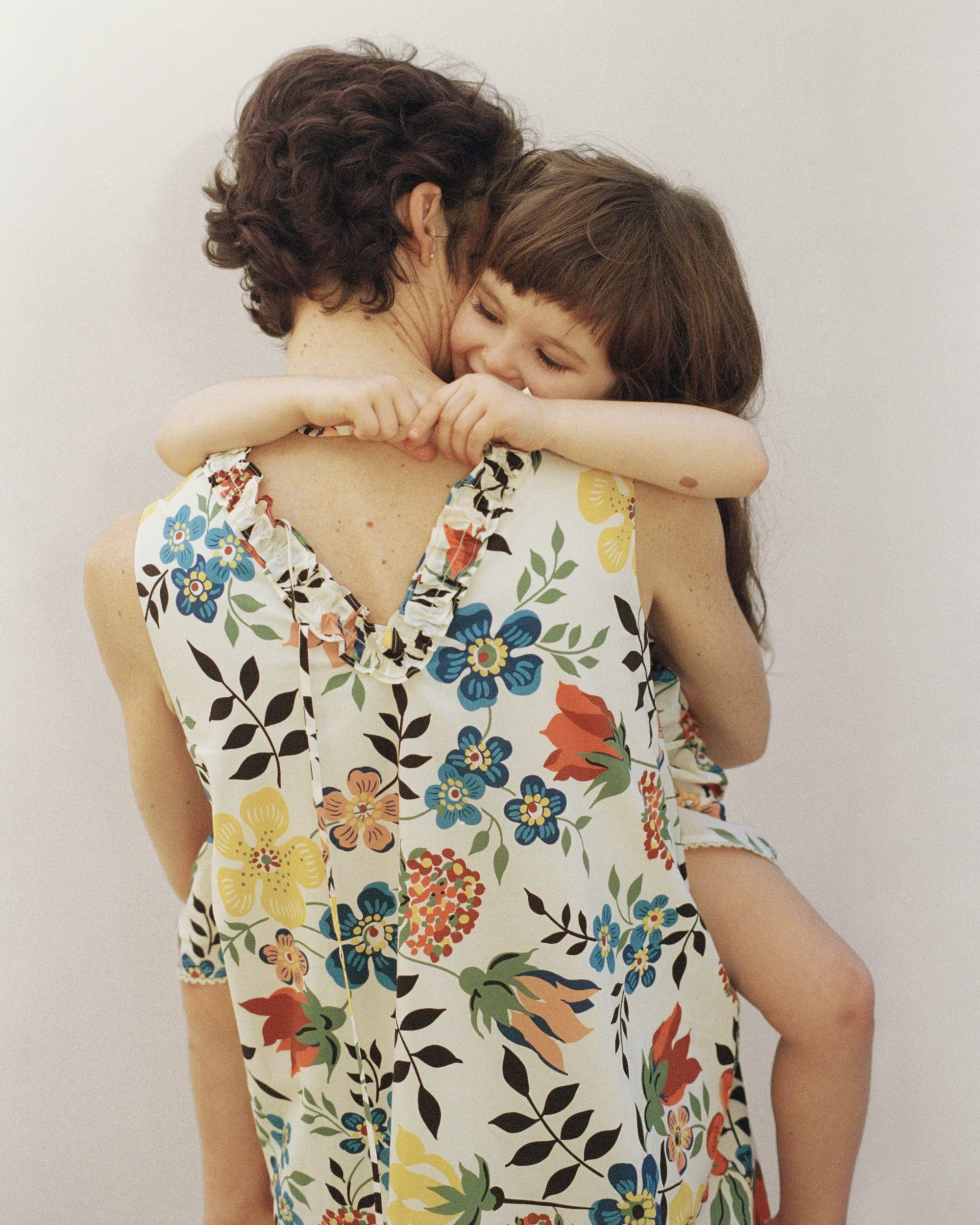 An adult and a child embracing warmly, photographed from behind; the adult wears a sleeveless V-back dress in lightweight fabric with a vibrant all-over Liberty floral print featuring blue, red, yellow and orange blooms with dark botanical stems on an ivory ground, with delicate ruffle trim at the neckline and hem — a fluid, relaxed silhouette against a soft neutral background