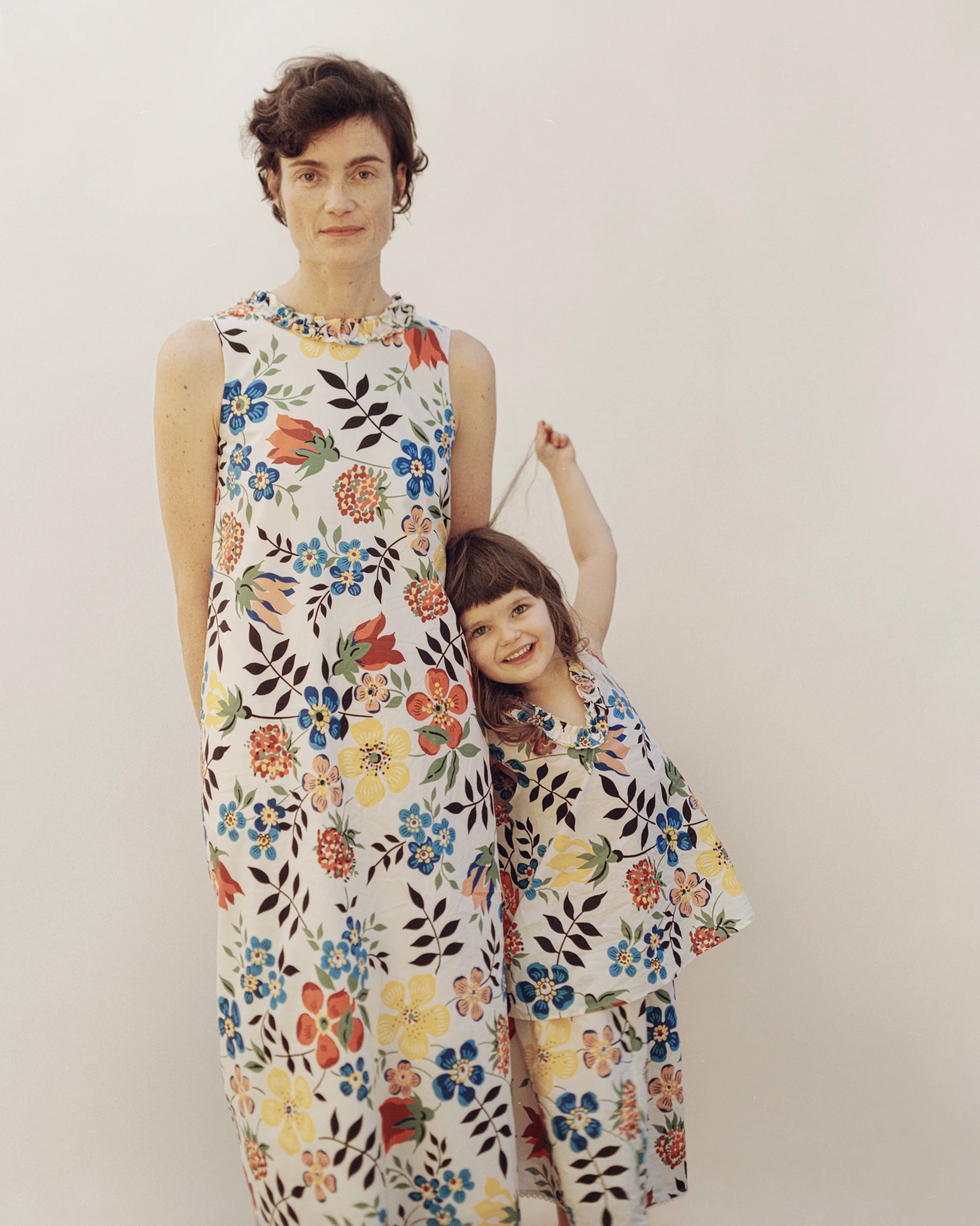 An adult woman and a young girl wearing matching sleeveless maxi dresses in crisp, lightweight fabric with a vibrant all-over Liberty floral print featuring blue, red, yellow and orange blooms with dark botanical stems on an ivory ground — the adult's dress has a ruffle crew neckline and a straight, column silhouette; the child wears a shorter version of the same print — both posed together against a soft neutral background