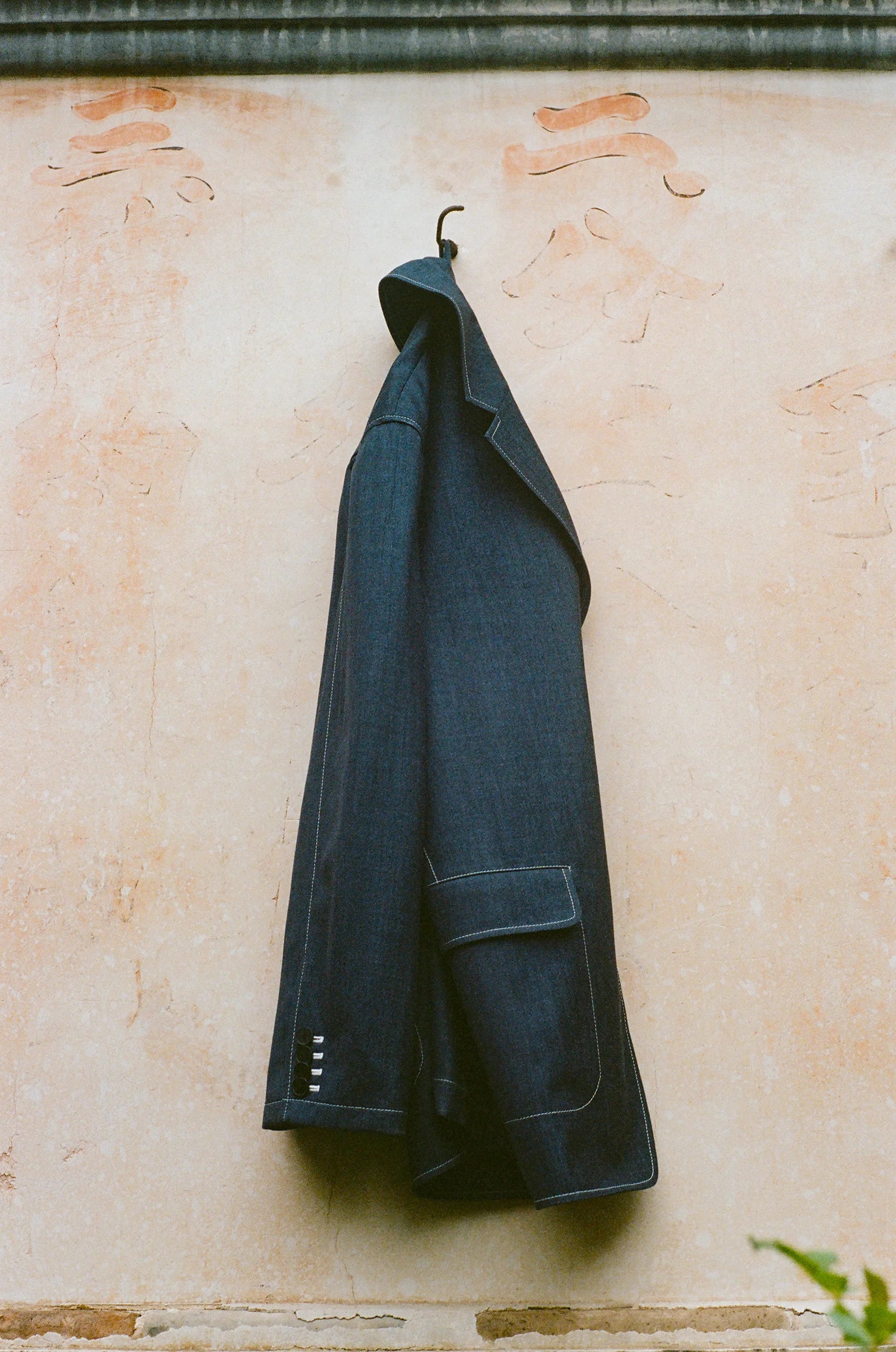 A indigo structured jacket hangs against a softly weathered wall, highlighting its texture and tailored construction details.