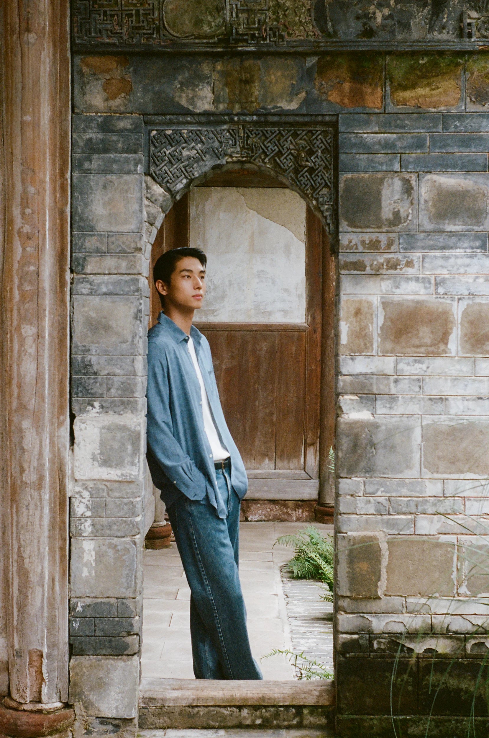 A model stands within the frame of an aged stone doorway, wearing relaxed blue denim and a lightweight shirt in a courtyard with traditional architecture.