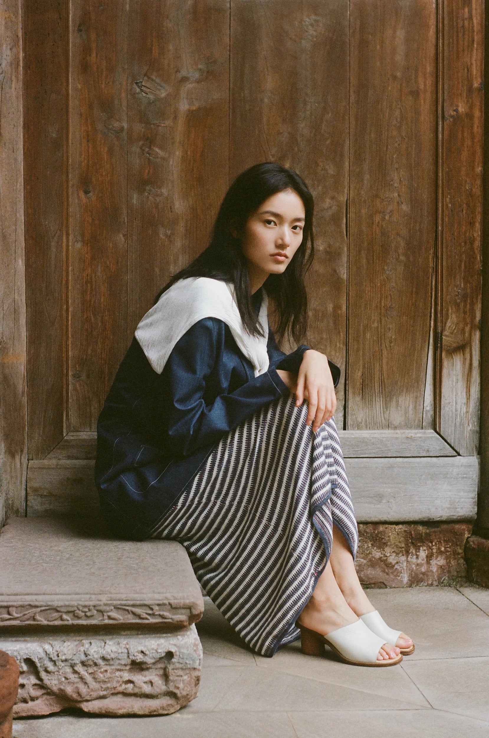 A model sits before a weathered wooden doorway, wearing a navy and ivory silhouette paired with a striped pleated skirt in a setting with traditional architecture.