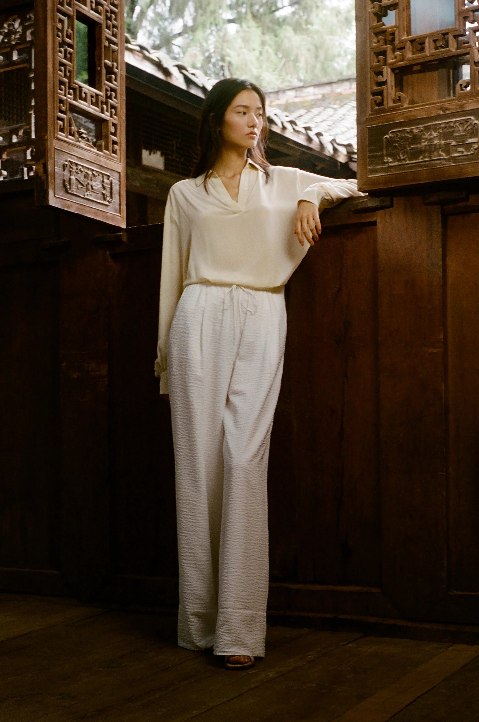 A model stands beside carved wooden windows, wearing a fluid ivory silhouette composed of a draped top and textured trousers within a traditional architectural interior.