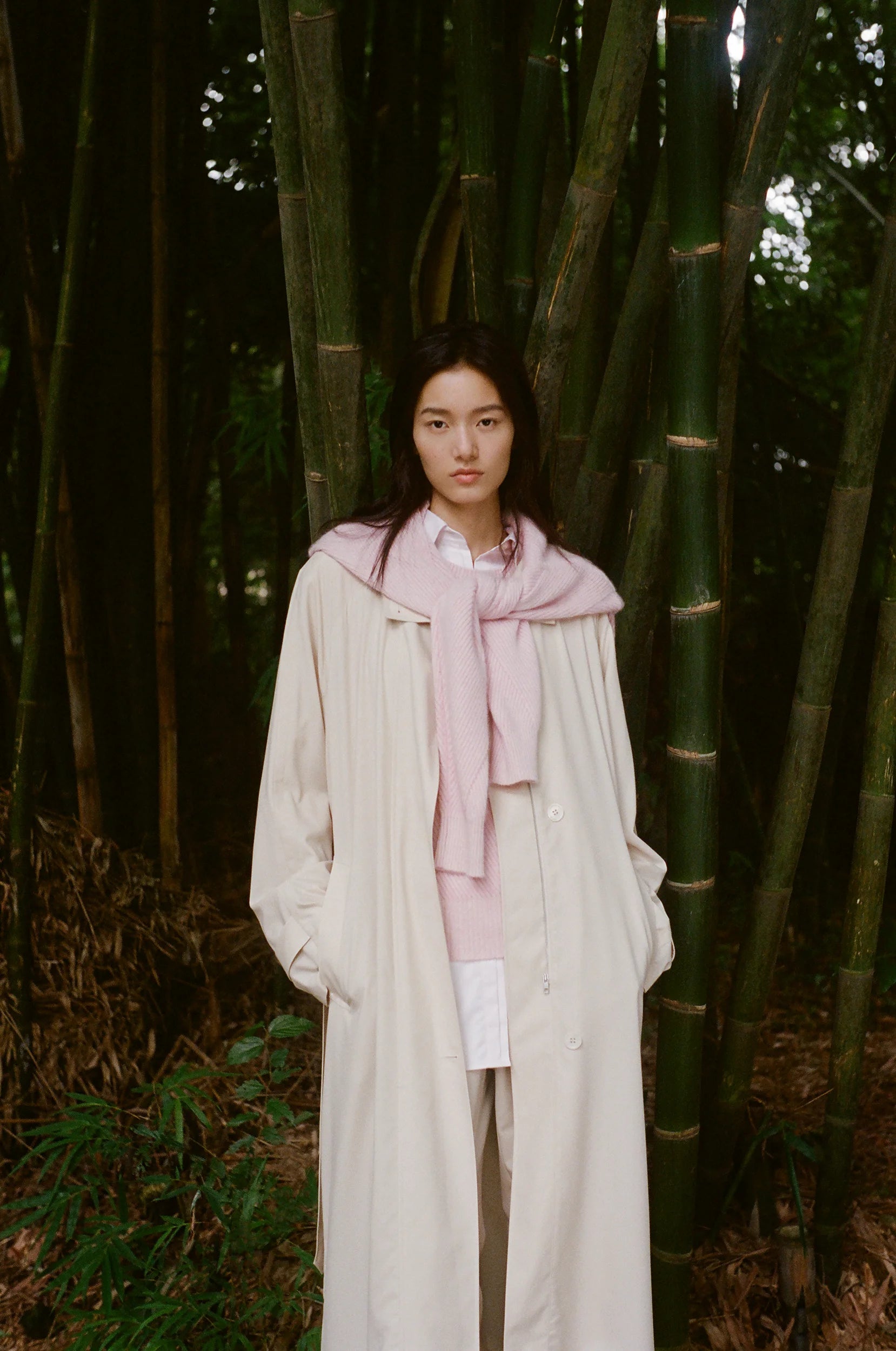 A model stands before a bamboo grove, wearing a flowing ivory trench coat layered over soft pale pink and white garments, in a calm and natural setting.