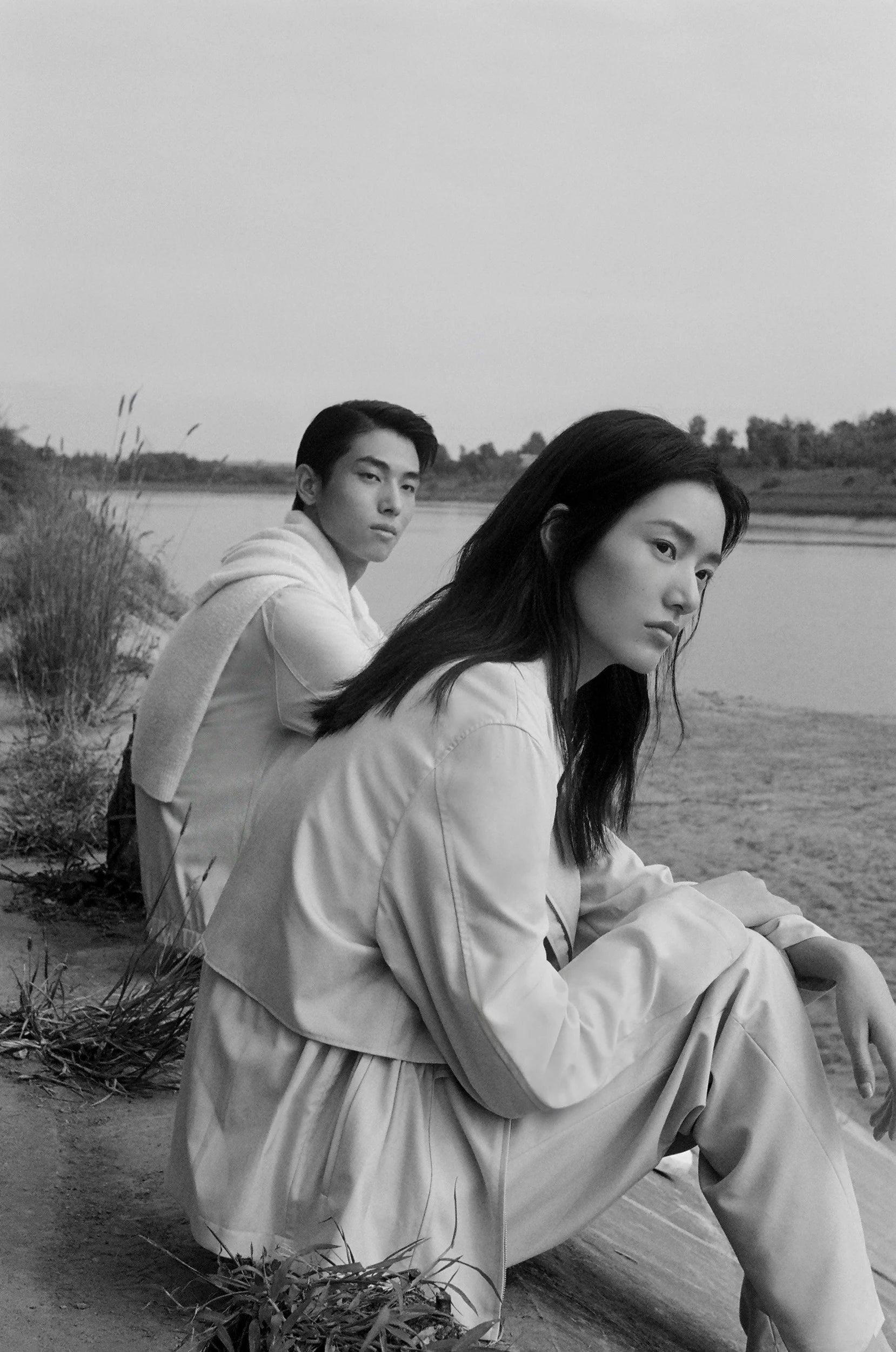 Two models seated by the water wear fluid light-toned silhouettes in a black-and-white image with a calm and contemplative atmosphere.