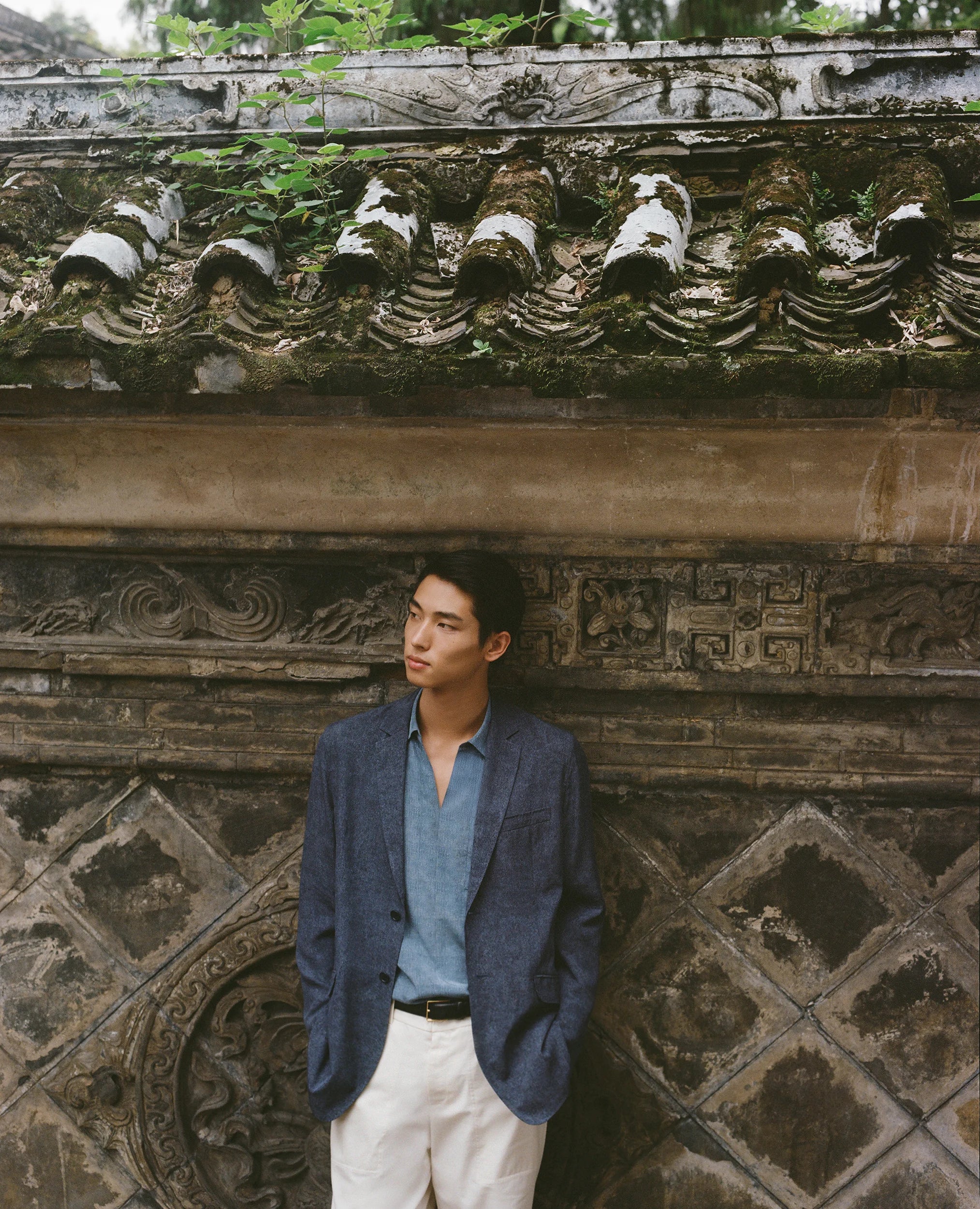 A model wearing a deep blue jacket, a light blue shirt, and ivory trousers stands before an aged stone wall with carved motifs and weathered vegetation.