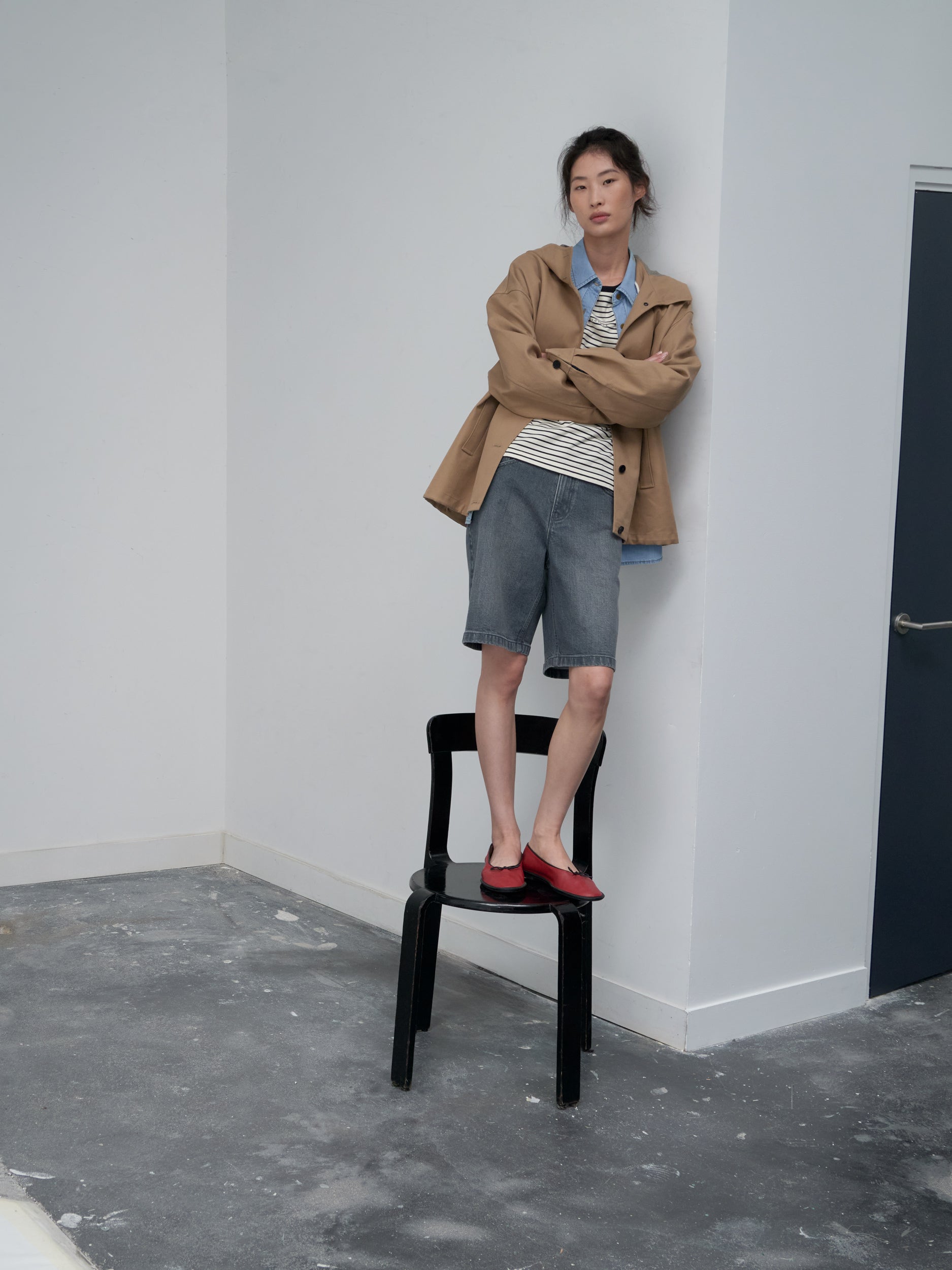 A woman stands on a black chair in a minimalist studio corner, wearing a camel jacket layered over a striped top and light blue shirt, paired with knee-length denim shorts and red ballet flats.