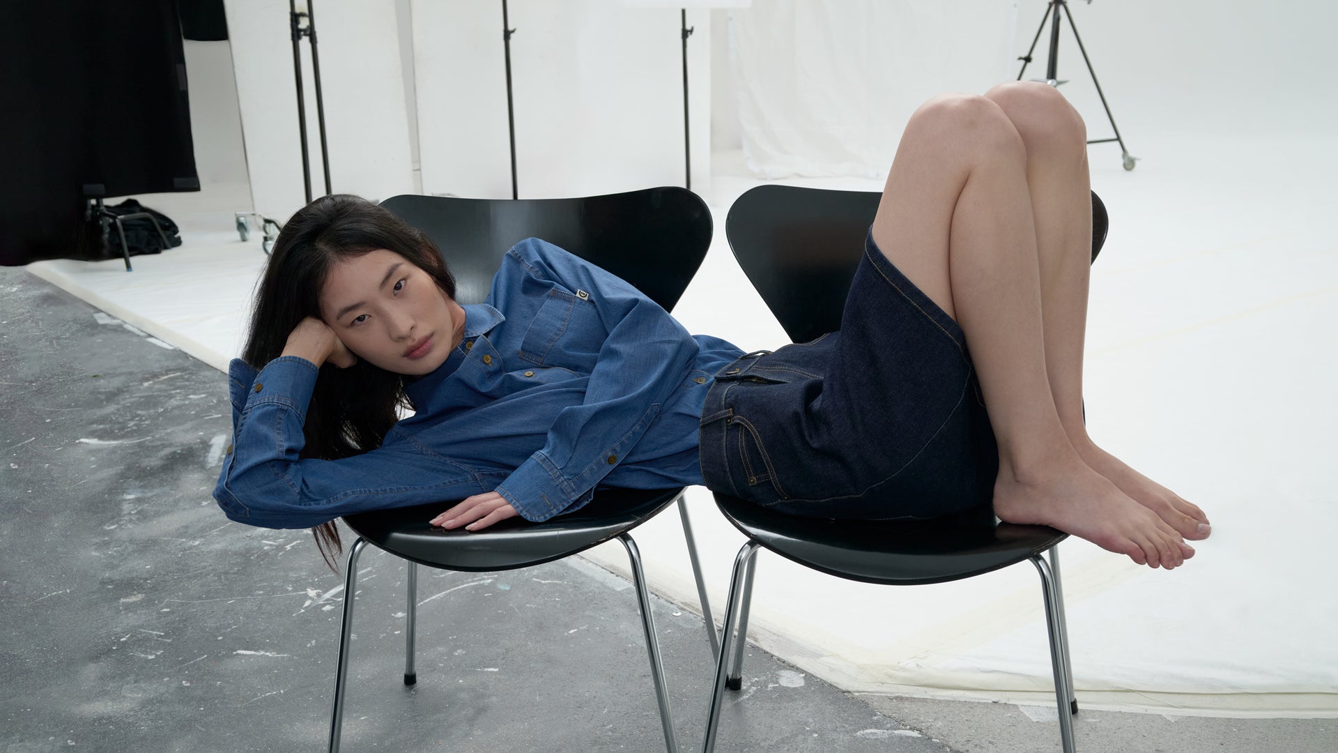 A model reclines across two black chairs in a studio setting, wearing a blue denim shirt with visible stitching and a dark indigo denim skirt. The look highlights a coordinated indigo ensemble with structured tailoring and contrast topstitch details.