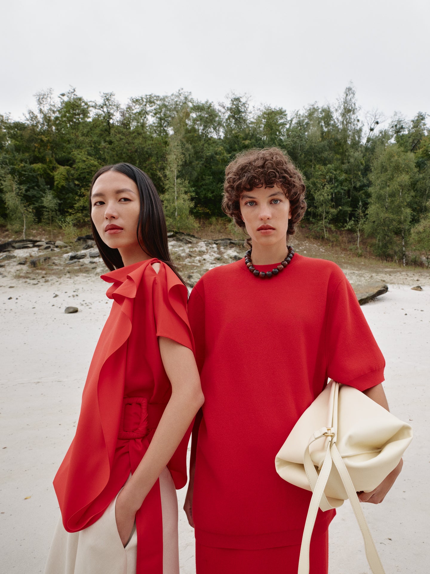 Two women stand side by side on a sandy landscape bordered by trees. One wears a red short-sleeved top in a linen and cashmere blend, featuring soft, fluid lines, paired with high-waisted cream trousers with a clean, tailored cut. The other wears a red silk blouse with a loose, asymmetrical drape and soft gathers, paired with a matching red skirt. Both silhouettes emphasize light fabrics, flowing volumes, and refined construction, contrasting with the natural setting.