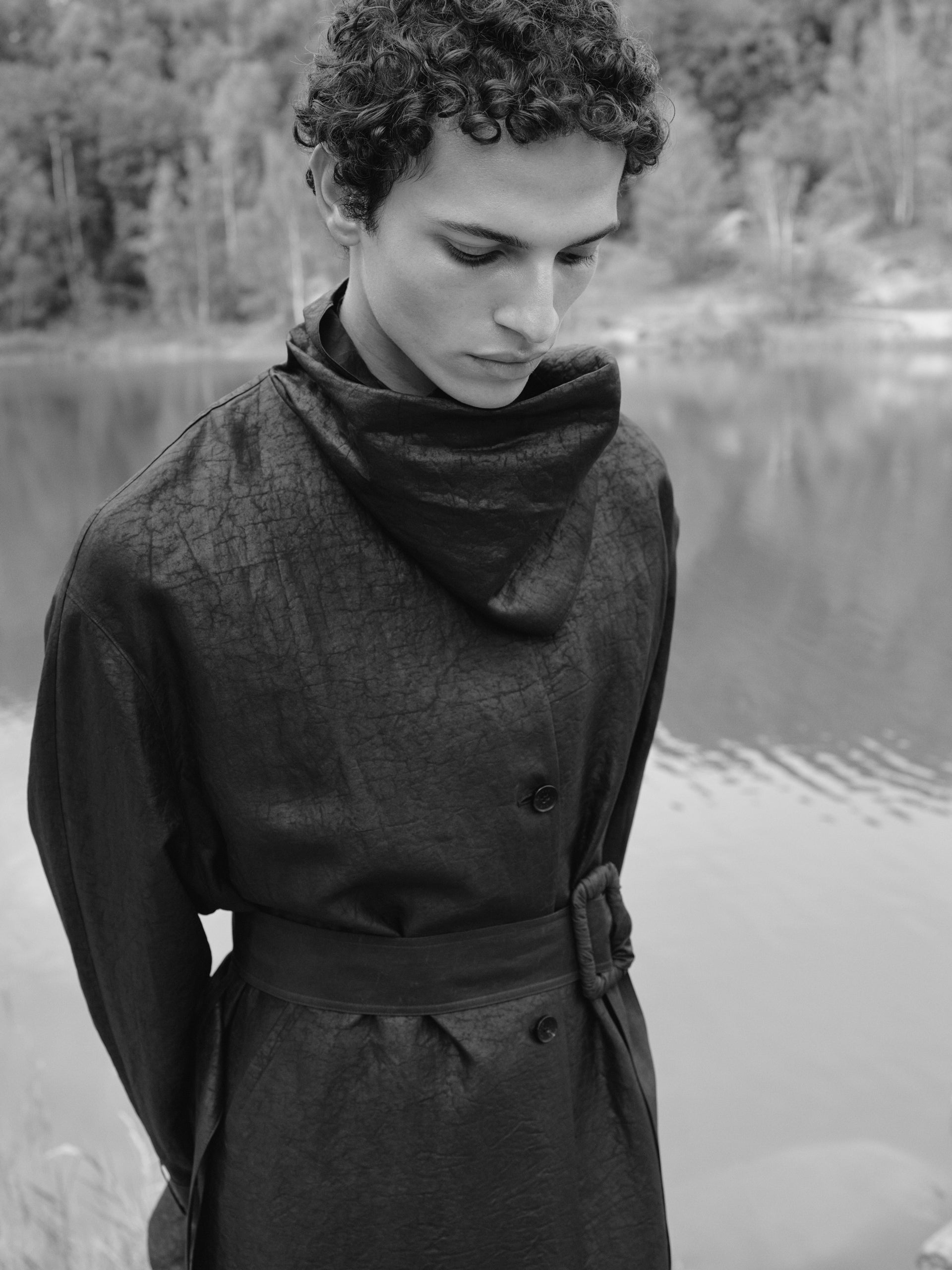 Black-and-white portrait of a man wearing a dark coat with a subtly silk crinkled texture, featuring a wrap-style collar and a tied belt at the waist. The silhouette is both structured and fluid. He stands by the water in a natural landscape with soft reflections, creating a calm, contemplative mood.