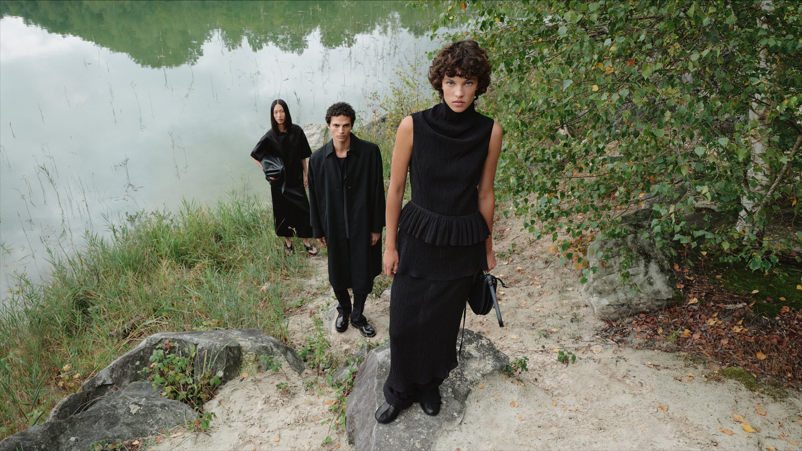Three people dressed in black express natural elegance. One woman wears a long silk dress with a high collar, a man wears a tailored suit, and another woman wears a black top paired with a skirt and hols a leather bag.