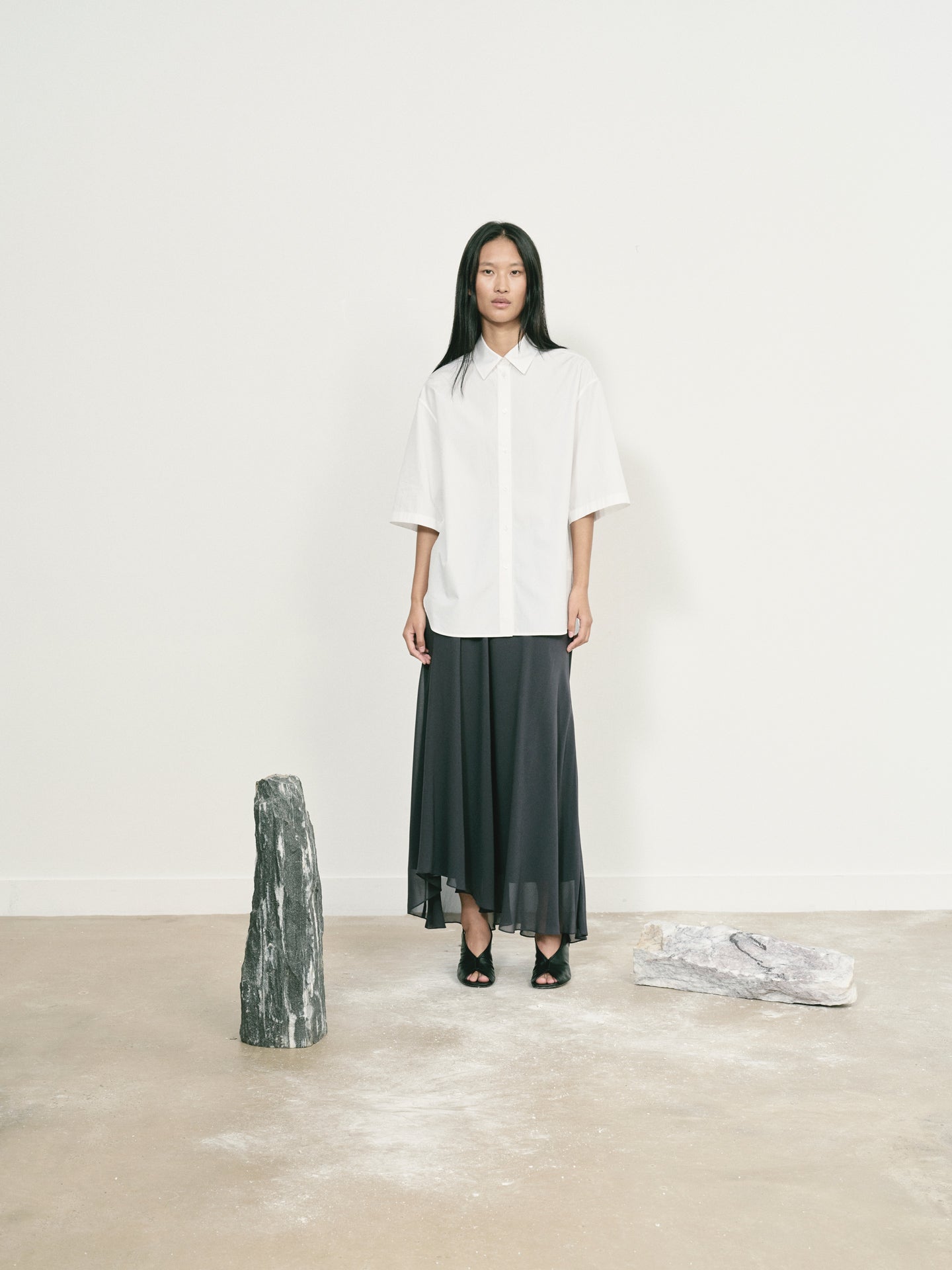 Person wearing a white shirt and dark skirt standing in a minimalistic setting with a white wall and beige floor.
