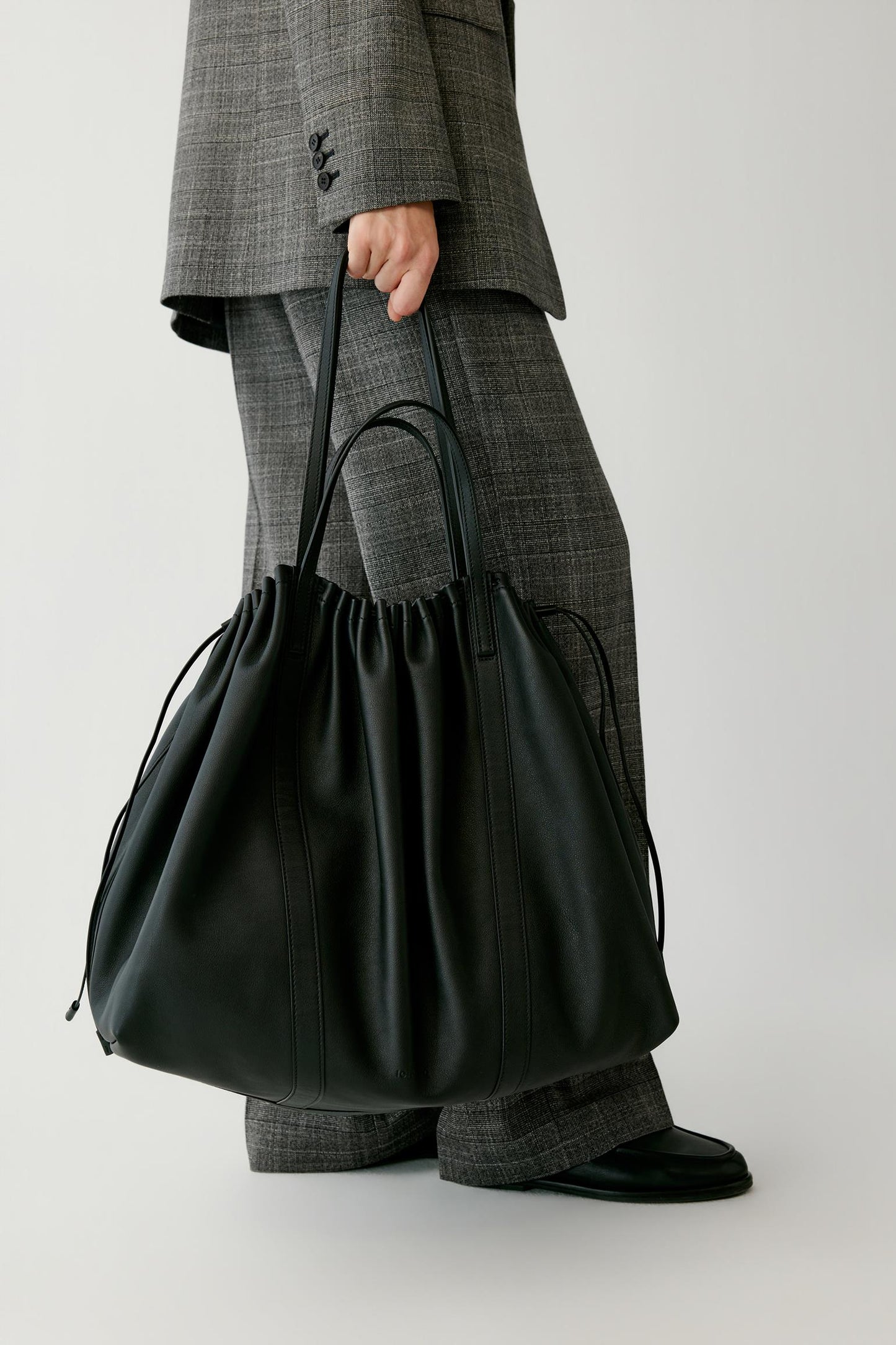 A person wearing a gray plaid suit holds a large black leather drawstring tote bag against a plain light gray studio background.