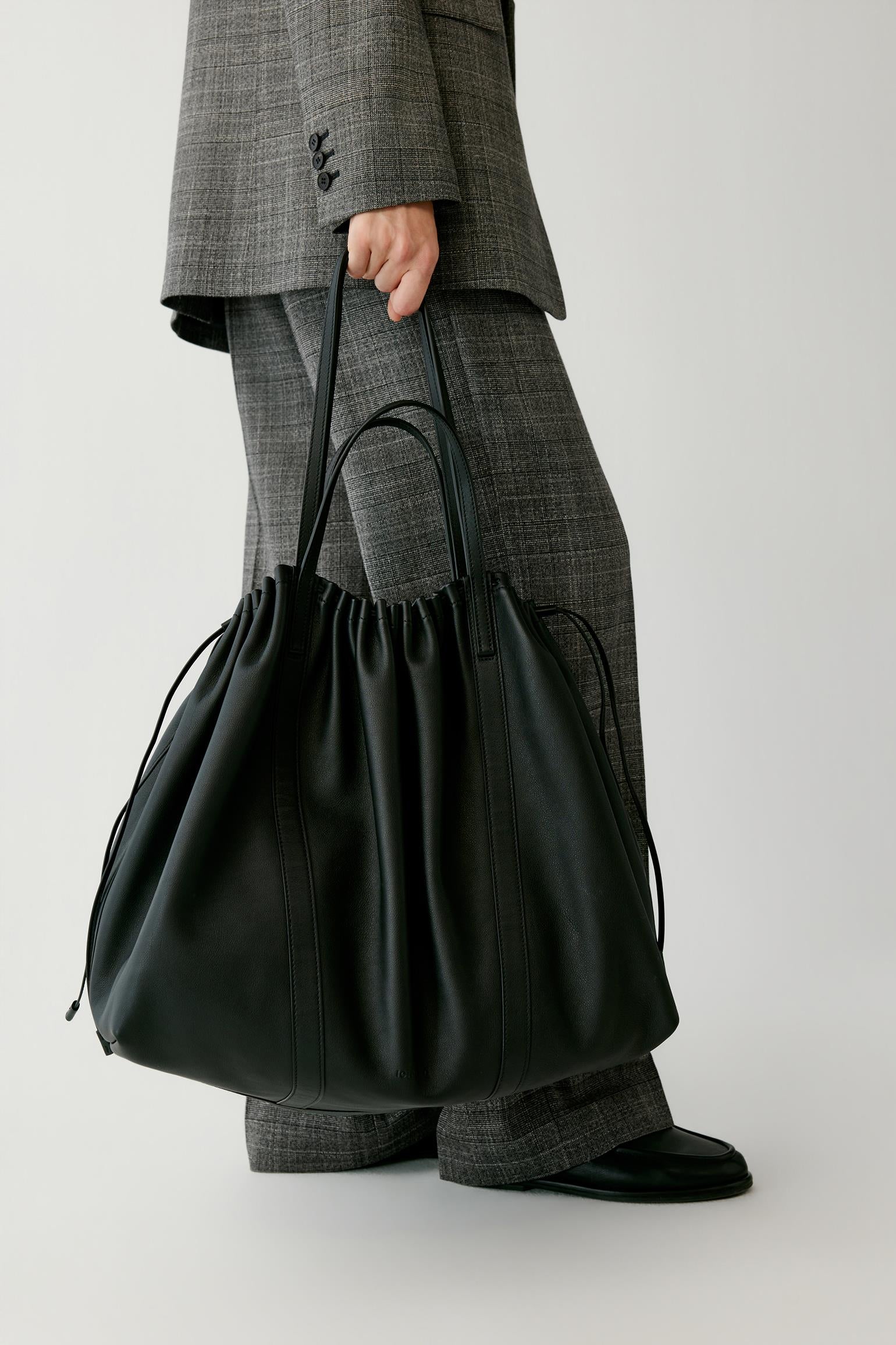 A person wearing a gray plaid suit holds a large black leather drawstring tote bag against a plain light gray studio background.