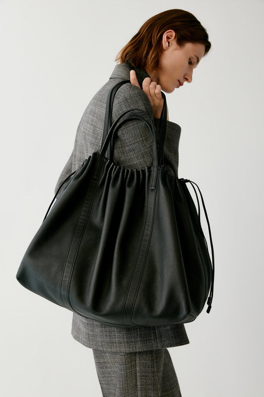 A woman in a gray plaid blazer and trousers carries a large black leather drawstring tote bag over her shoulder, photographed against a plain light gray studio background.