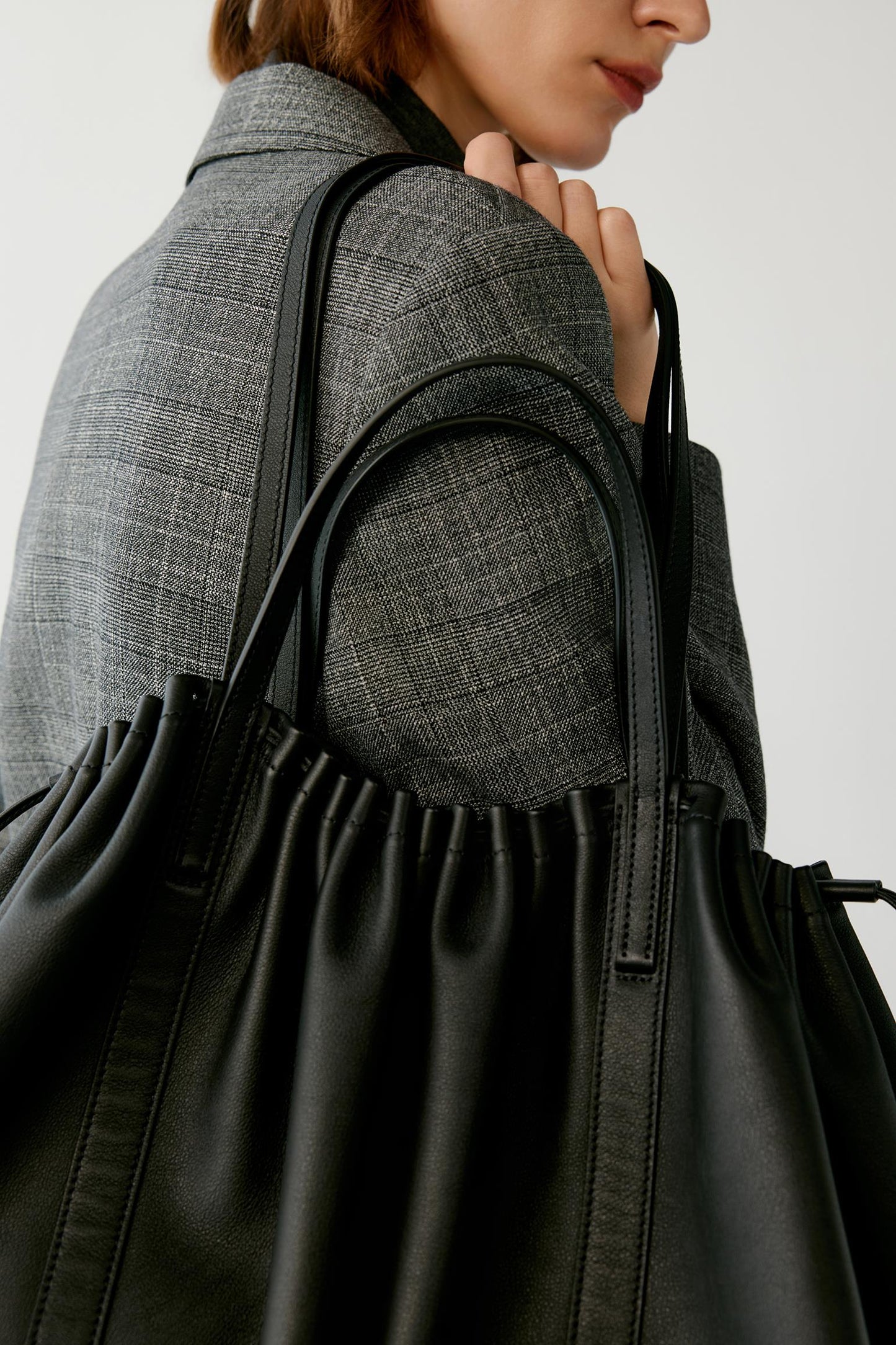 A close-up of a person wearing a gray textured blazer and carrying a large black leather tote bag with gathered detailing and double shoulder straps.
