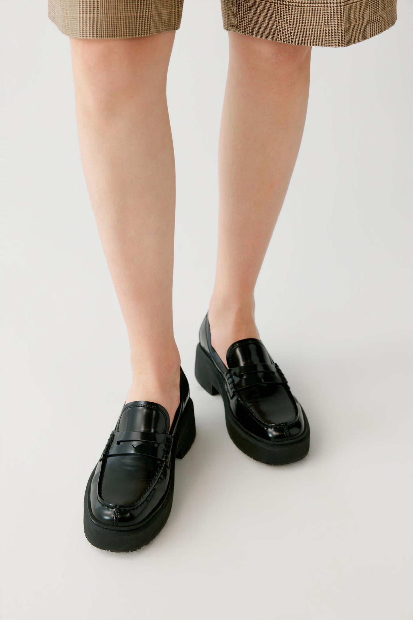 A person wearing black patent leather penny loafers with chunky platform soles, shown from the knees down against a light gray studio background.