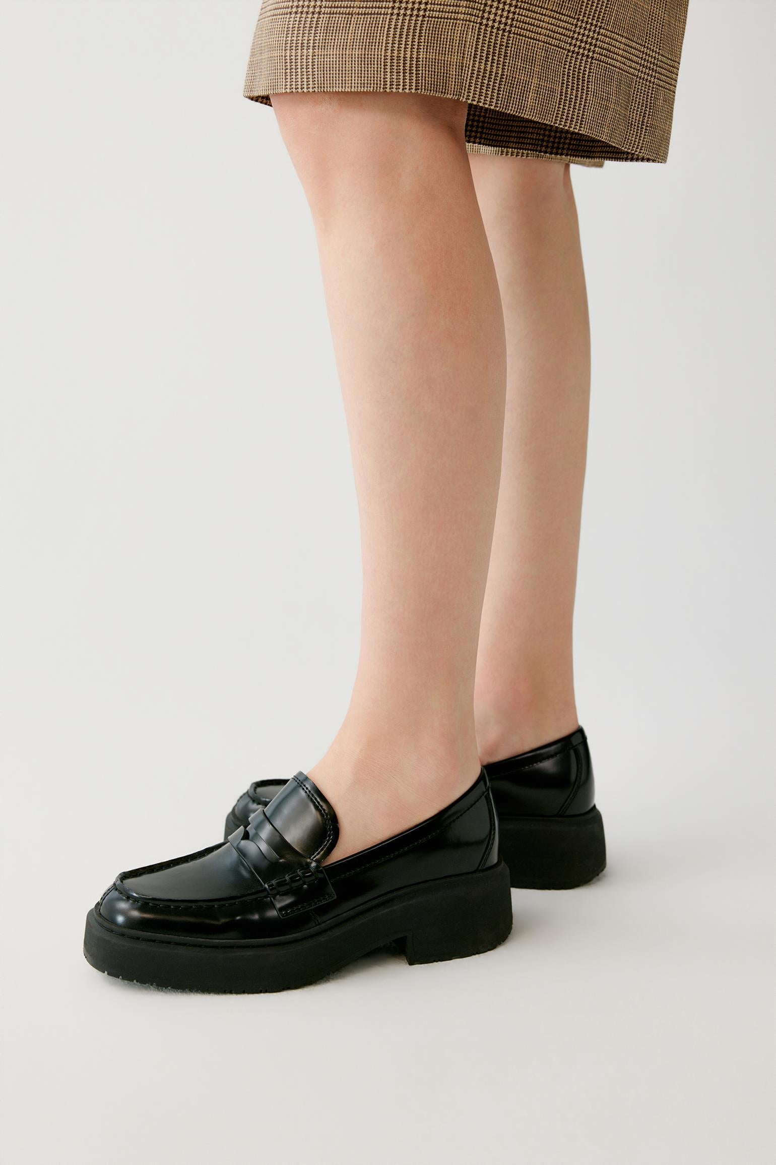 A person wearing black patent leather penny loafers with chunky platform soles, paired with a brown plaid skirt, shown from the knees down on a light gray studio background.