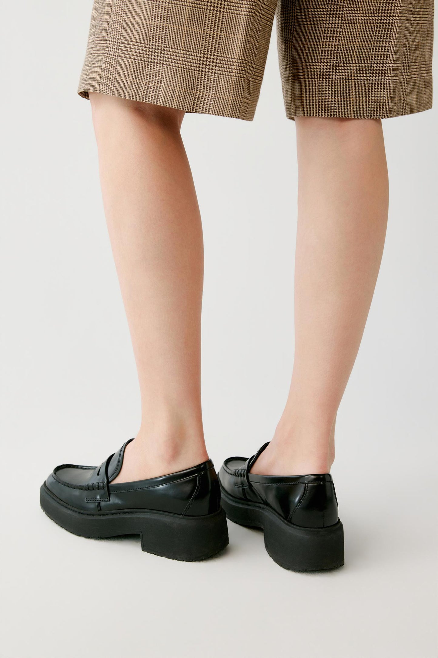 Black leather platform loafers worn with brown plaid shorts, shown from a rear three-quarter angle against a light gray studio background.