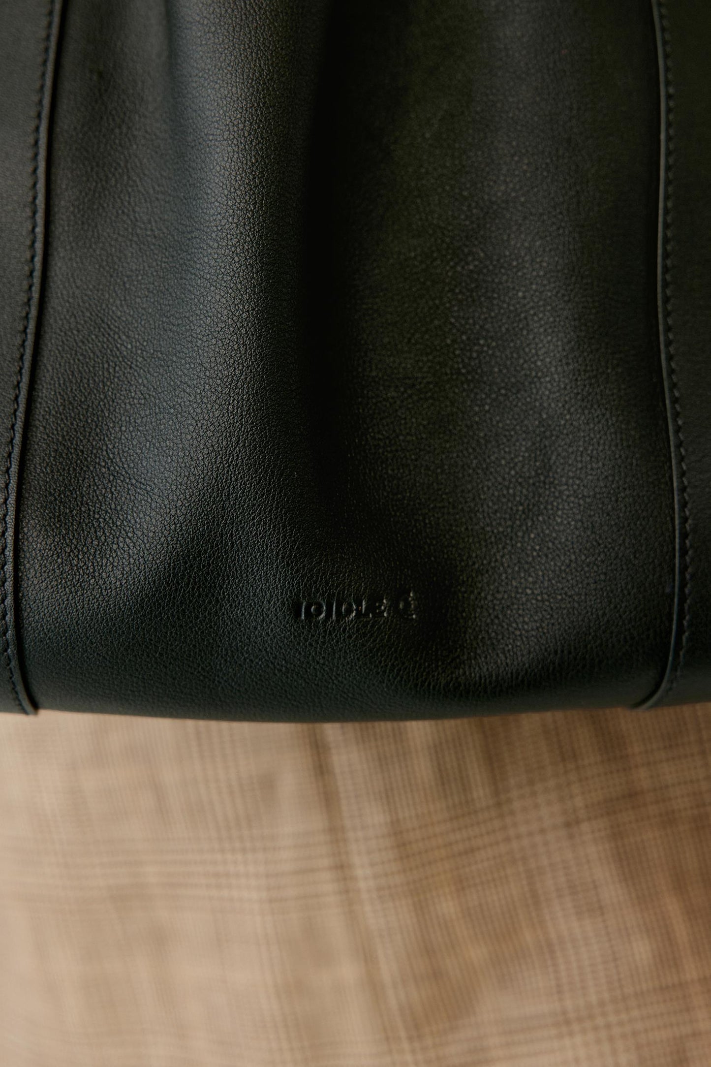 Close-up of a black leather tote bag with a debossed logo, resting on a beige plaid fabric surface.