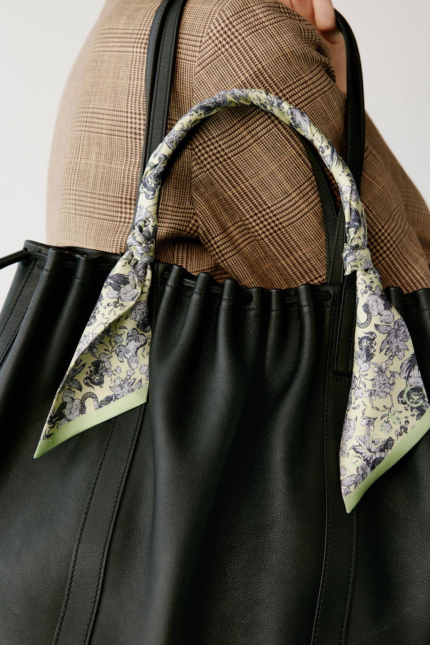 A close-up of a black leather tote bag with a decorative floral scarf tied to its handles, worn by a person in a brown plaid blazer.