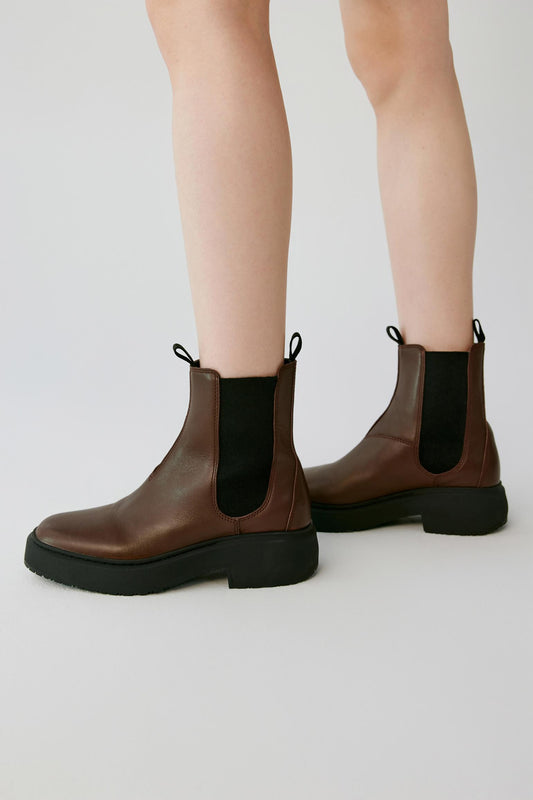 A pair of brown leather Chelsea boots with black elastic side panels and black soles, shown on a model's feet against a light gray studio background.