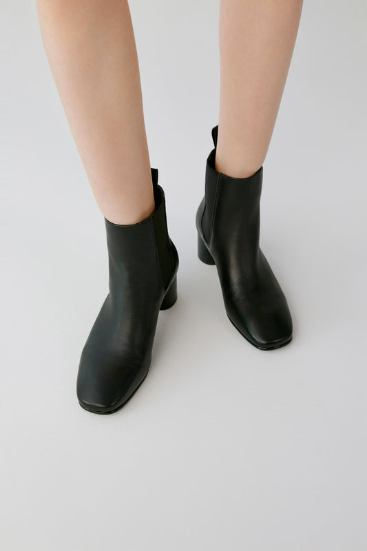 A pair of black leather ankle boots with square toes and low block heels, shown on a model's feet against a light gray studio background.