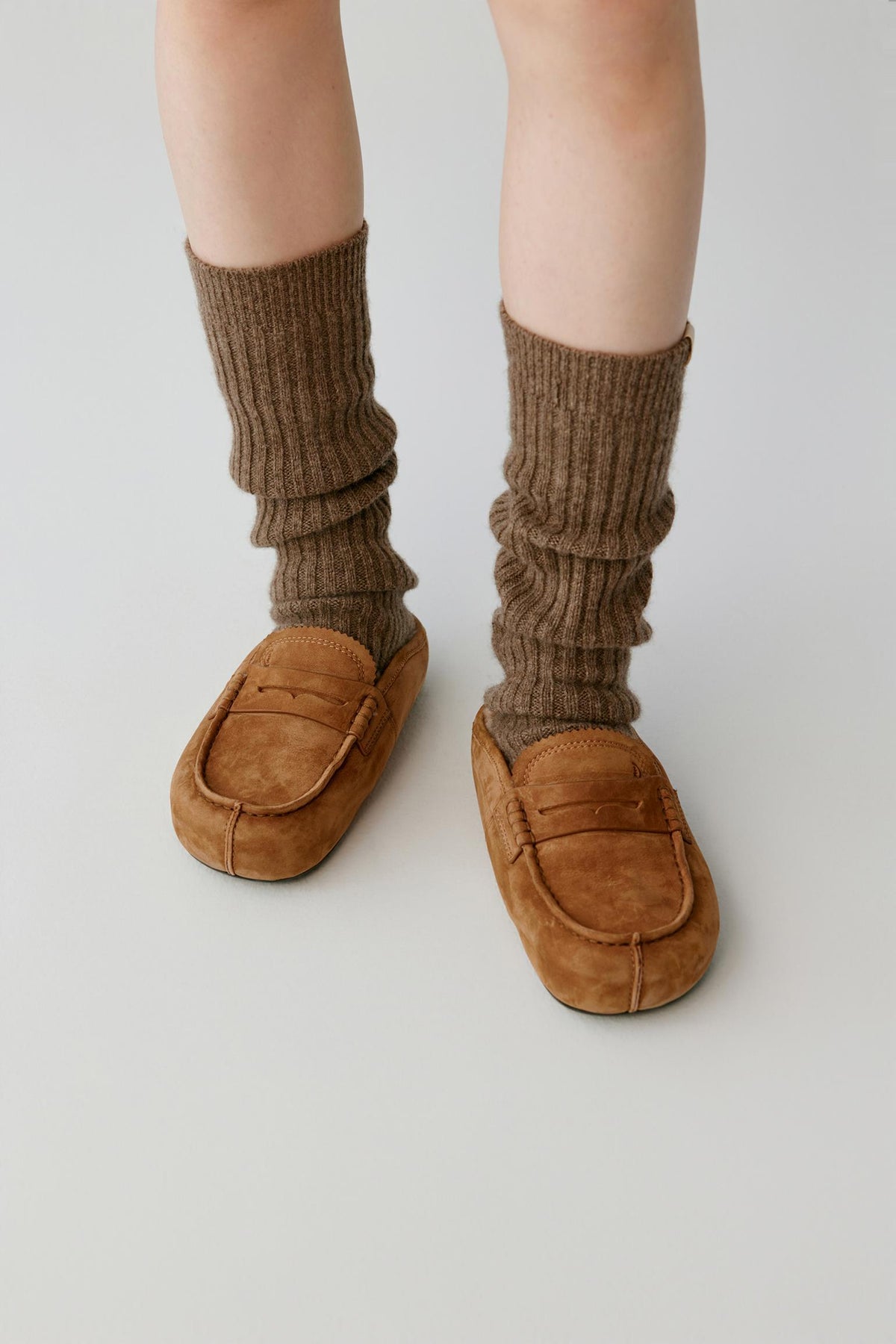 Front view of a woman wearing chestnut brown nubuck mules, showing the rounded toe, penny-loafer strap and stitched detailing.