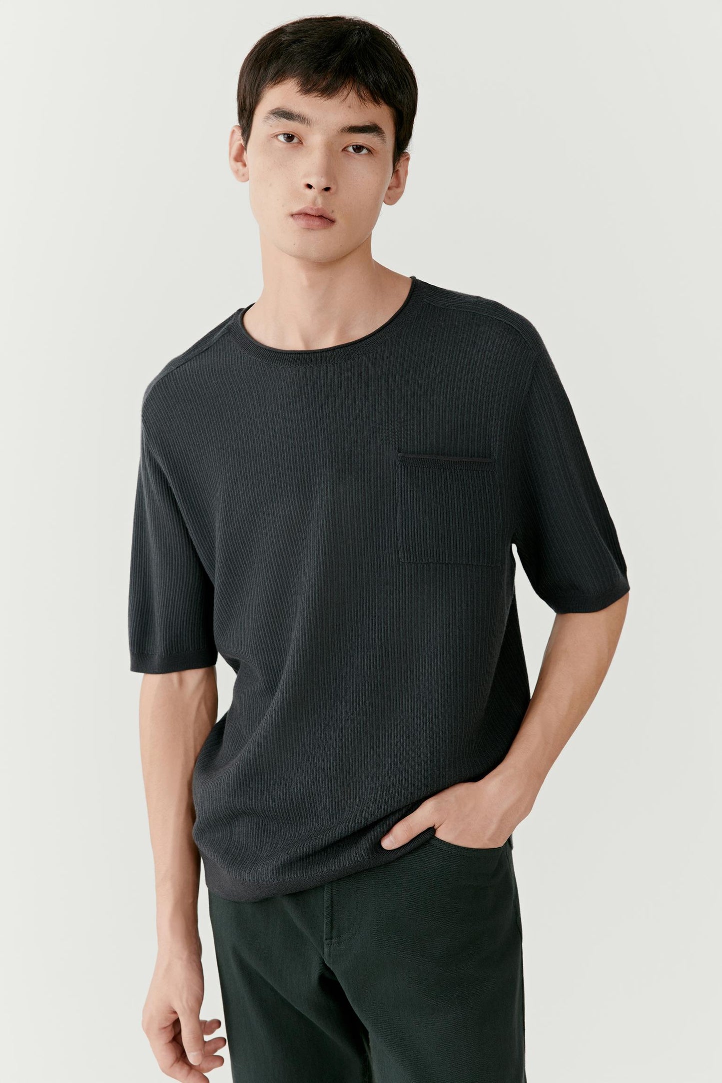 Man wearing a dark green wool T-shirt with a straight, relaxed fit, mid-length front view showing the soft knit texture, clean round neckline and short sleeves, one hand in pocket.